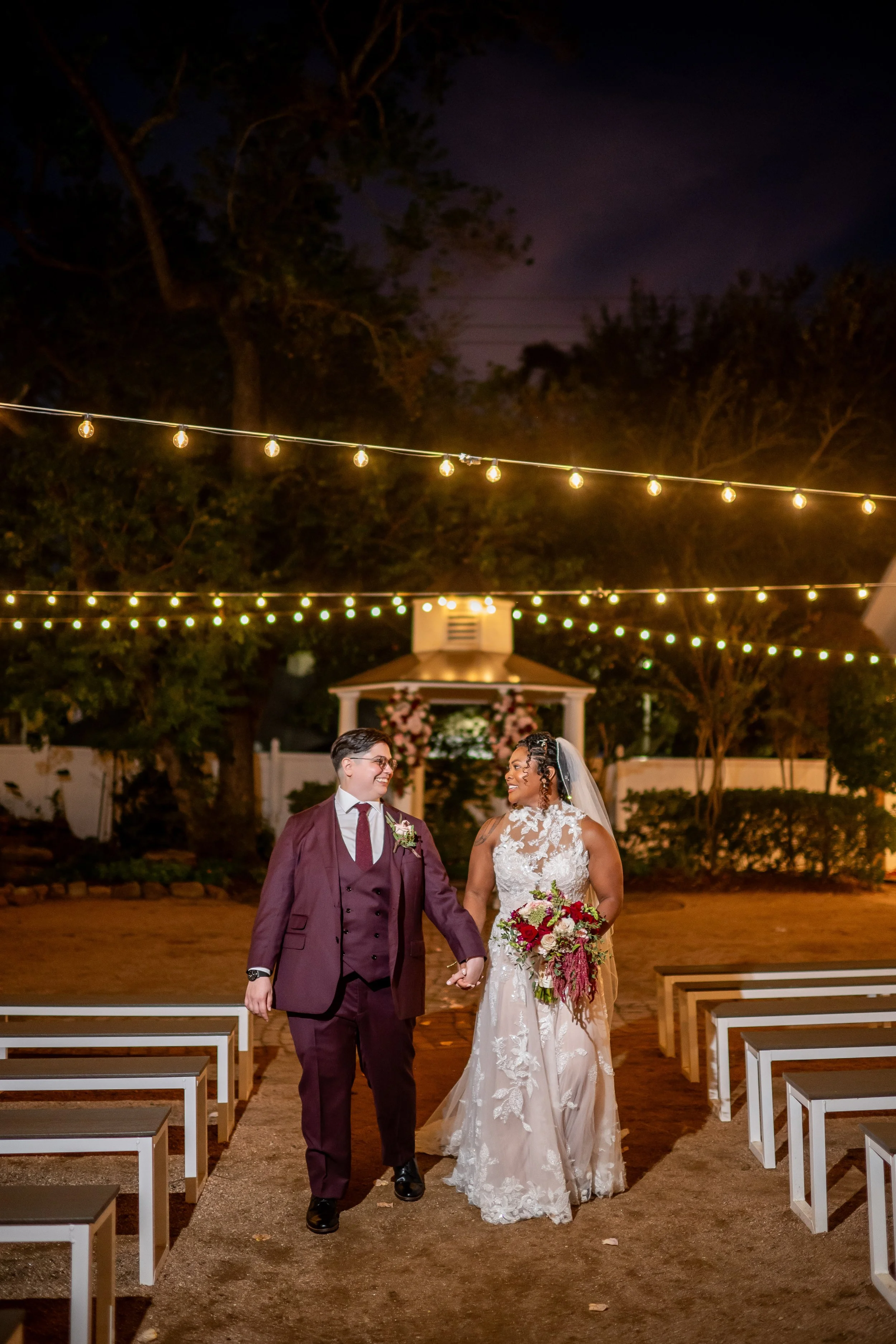 LGBTQ+ newlyweds hold hands while walking down an outdoor ceremony aisle under string lights.