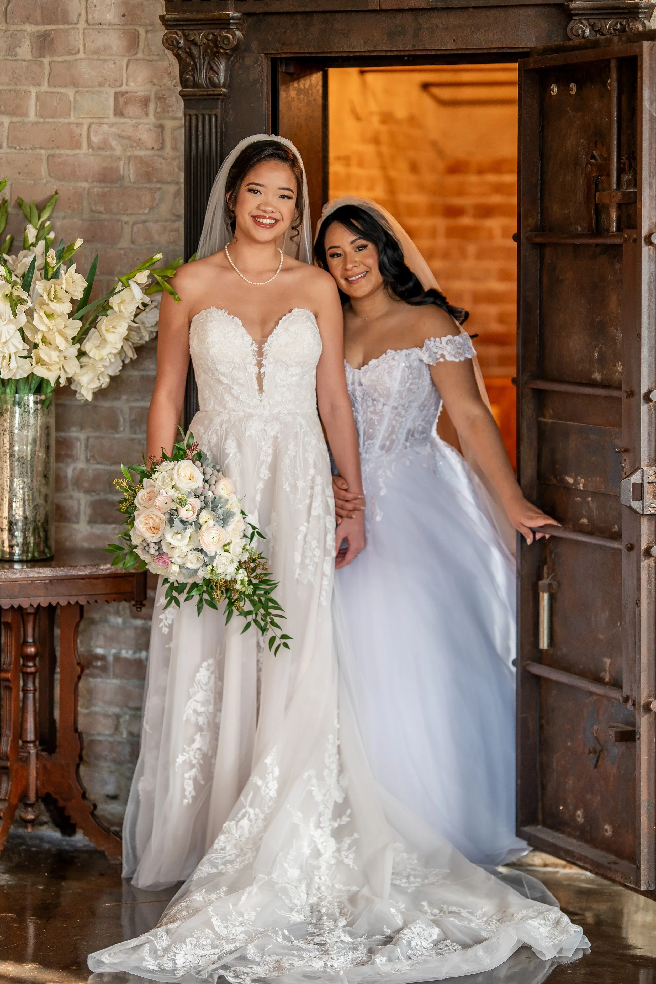 Two brides in wedding gowns stand together beside the historic vault at Butler’s Courtyard.