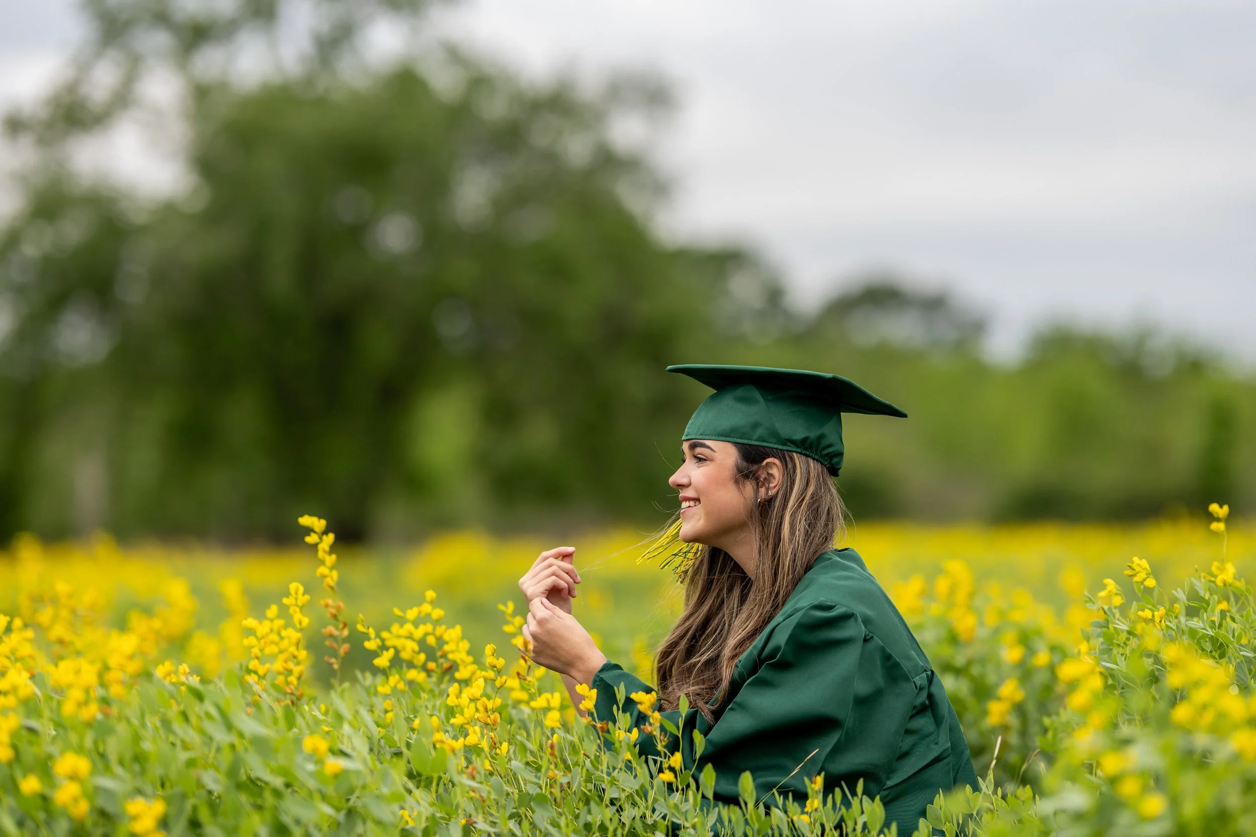 Graduate wearing a green cap and gown seated in a field of yellow wildflowers for a senior portrait session.