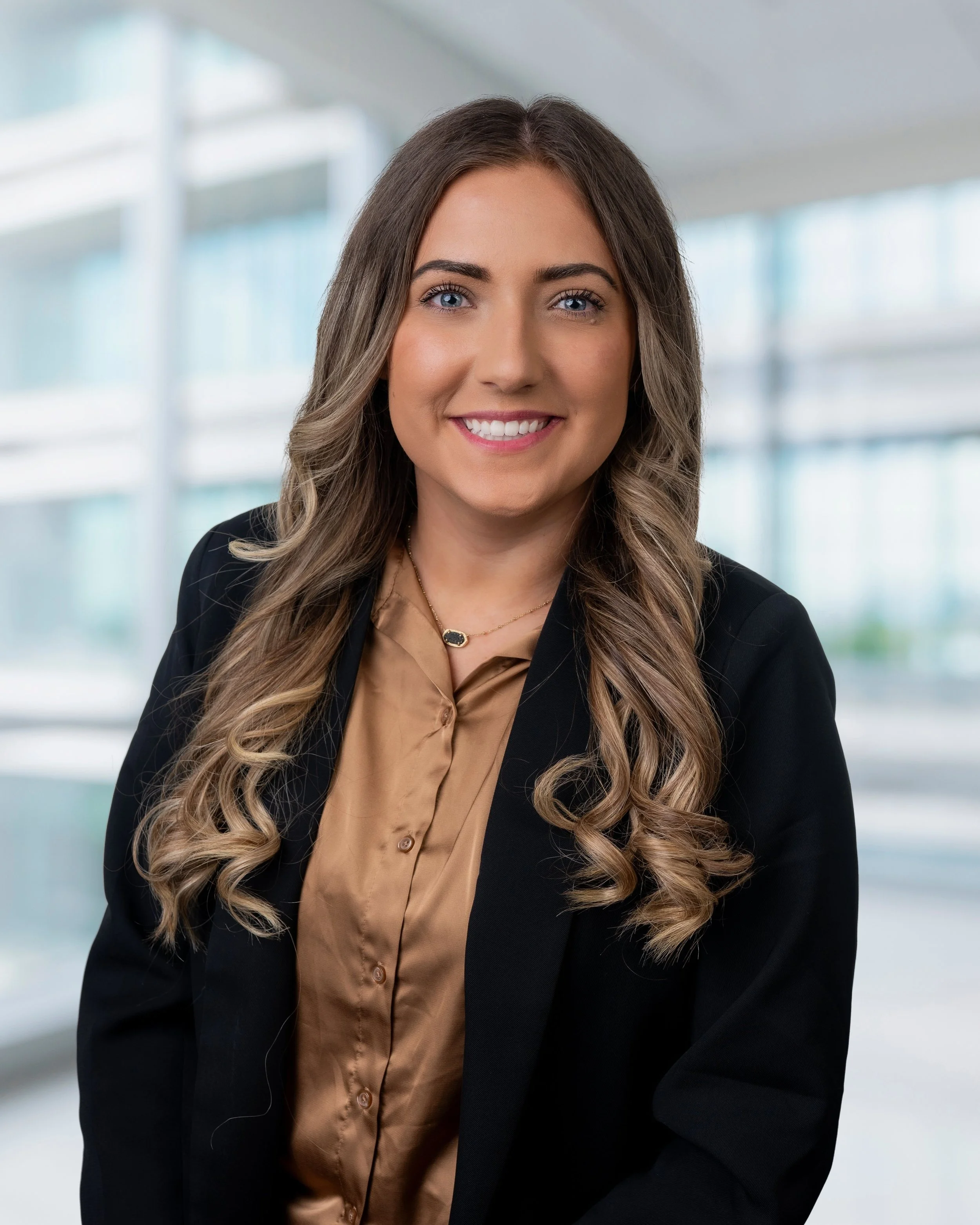 Professional business headshot of a smiling woman wearing a black blazer in a bright office setting.