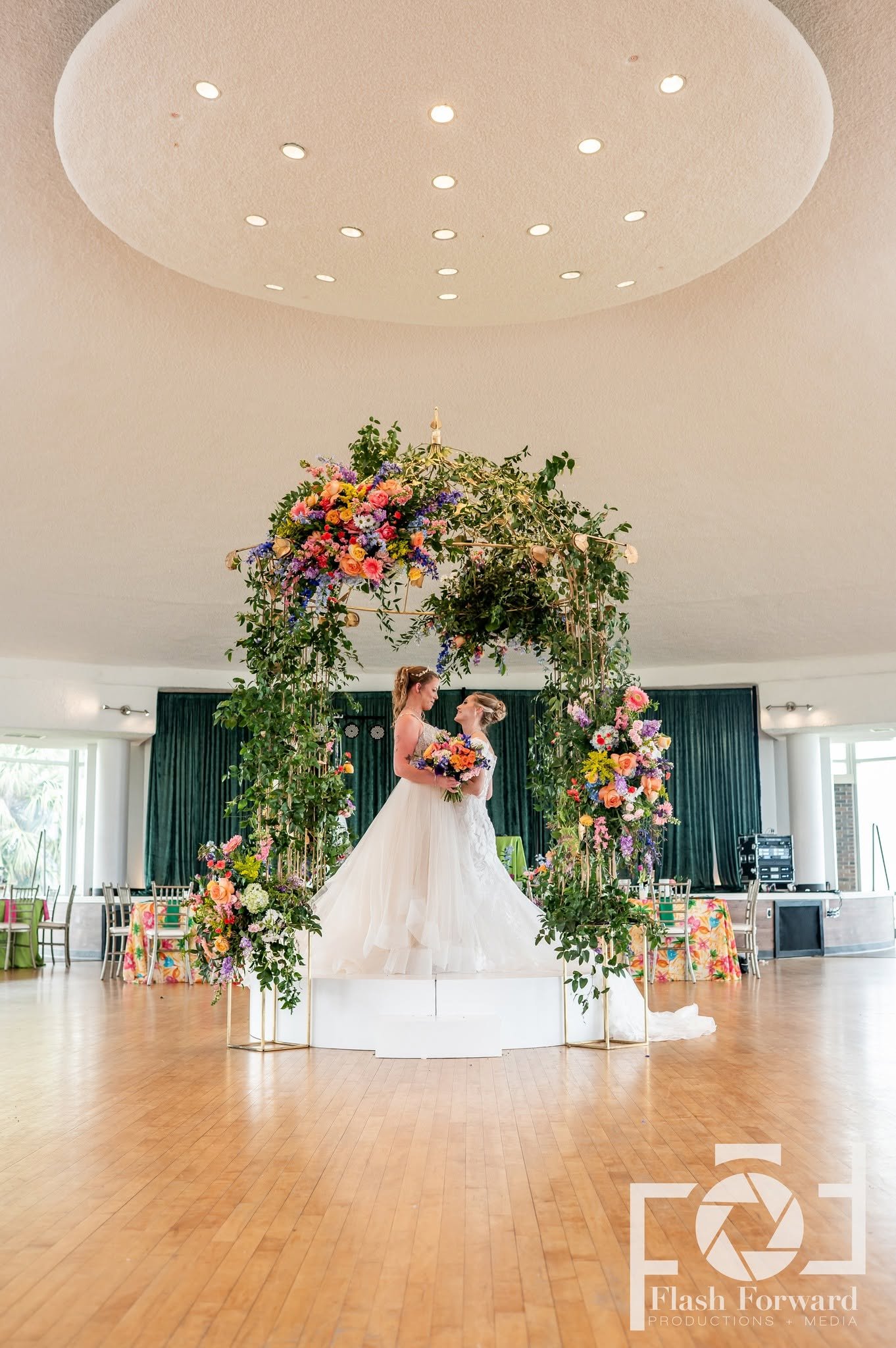 Two brides stand together beneath a large arch covered in colorful flowers and greenery.