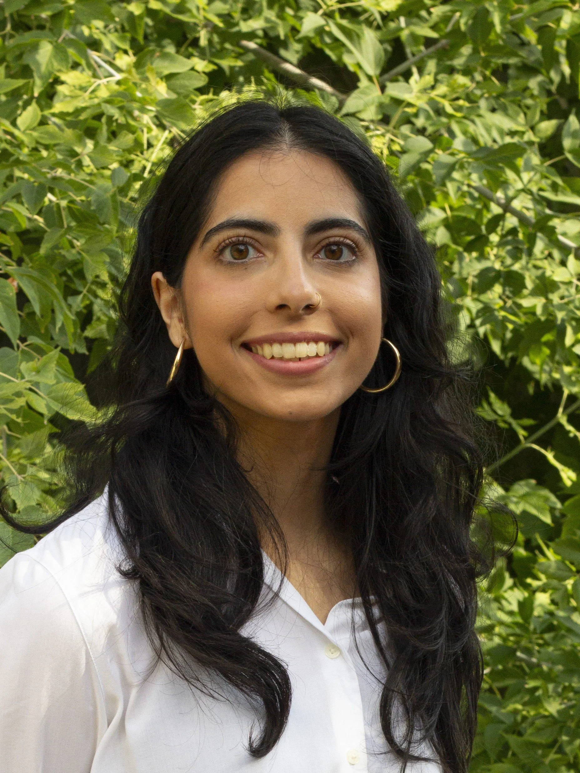 Aqsa Hussain stands before green bushes. She is smiling and has dark hair and wears a white blouse.