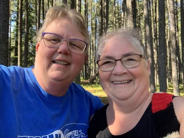 Two smiling women take a selfie in front of a forest of trees.