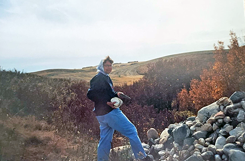 A woman in jeans, sweater and a kerchief stands over a stone pile. She is holding stones in her hands and smiling.