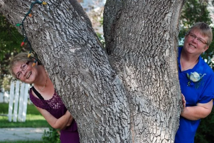 Two women are playfully peeking out from either side of a large tree. One has a blue shirt with a white rose boutonniere, the the other has a knitted dark purple top.