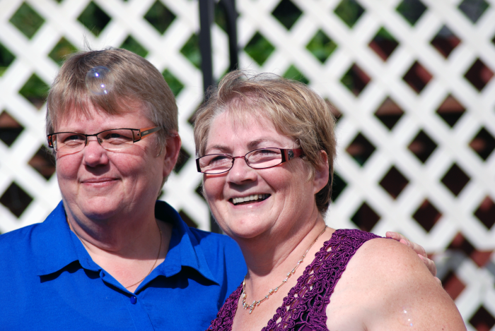 Two smiling women stand in front of a white fence. There are bubbles floating in the the air around them.