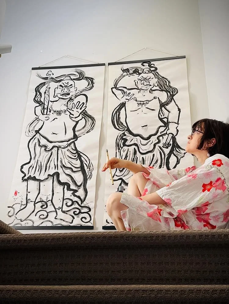 These two Kongōrikishi (Niō) statues are inspired by the concept of yin and yang. Chikako Ogawa sits in front of them, holding a paintbush and wearing a white dress with pink flowers.