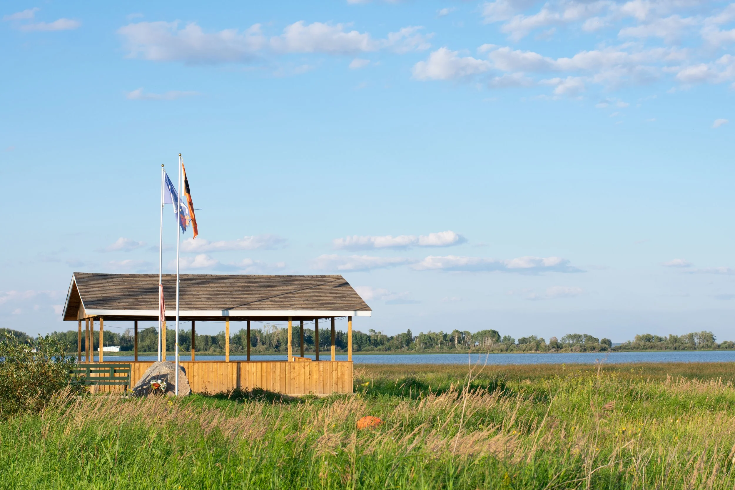 An open wooden picnic shelter overlooks a blue and reedy wetland under a blue sky with scattered clouds. Two flags are raised next to it.