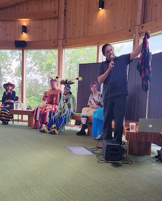 A man in a black shirt and pants hold up a brightly colored floral scarf. He is talking into a microphone. Several youth in Indigenous regalia are seated on benches to his right.