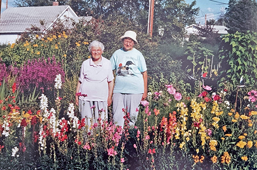Two women stand smiling in a lush and colorful garden. They are wearing t-shirts and sweat pants.
