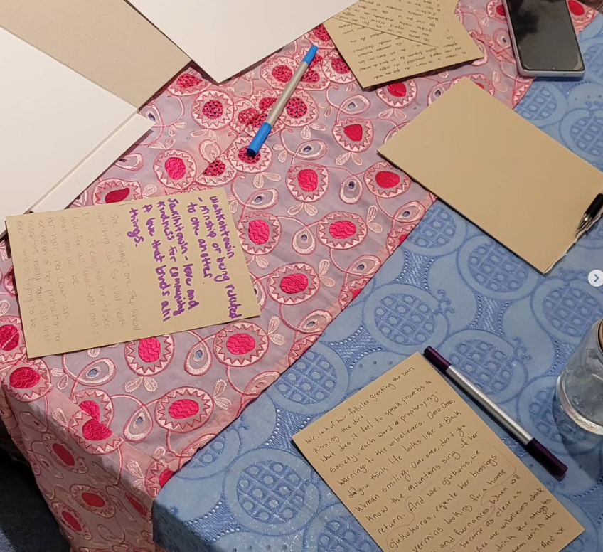 Colourful textiles spread across a table. Pens and pages with writing on them are scattered over the fabric.
