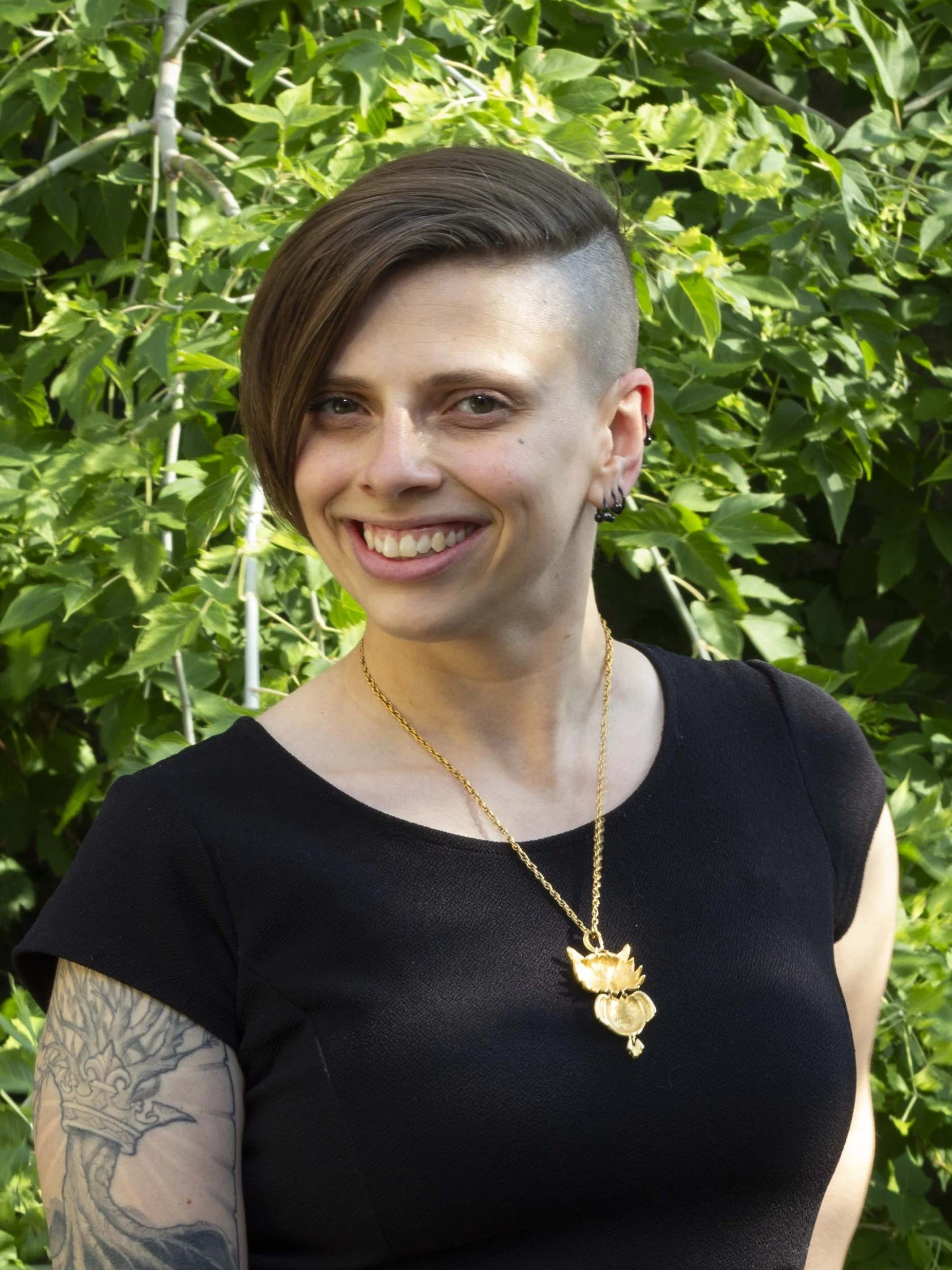 Jessica Jack stands in front of green bushes. She is smiling and wears a black t-shirt and a gold necklace.