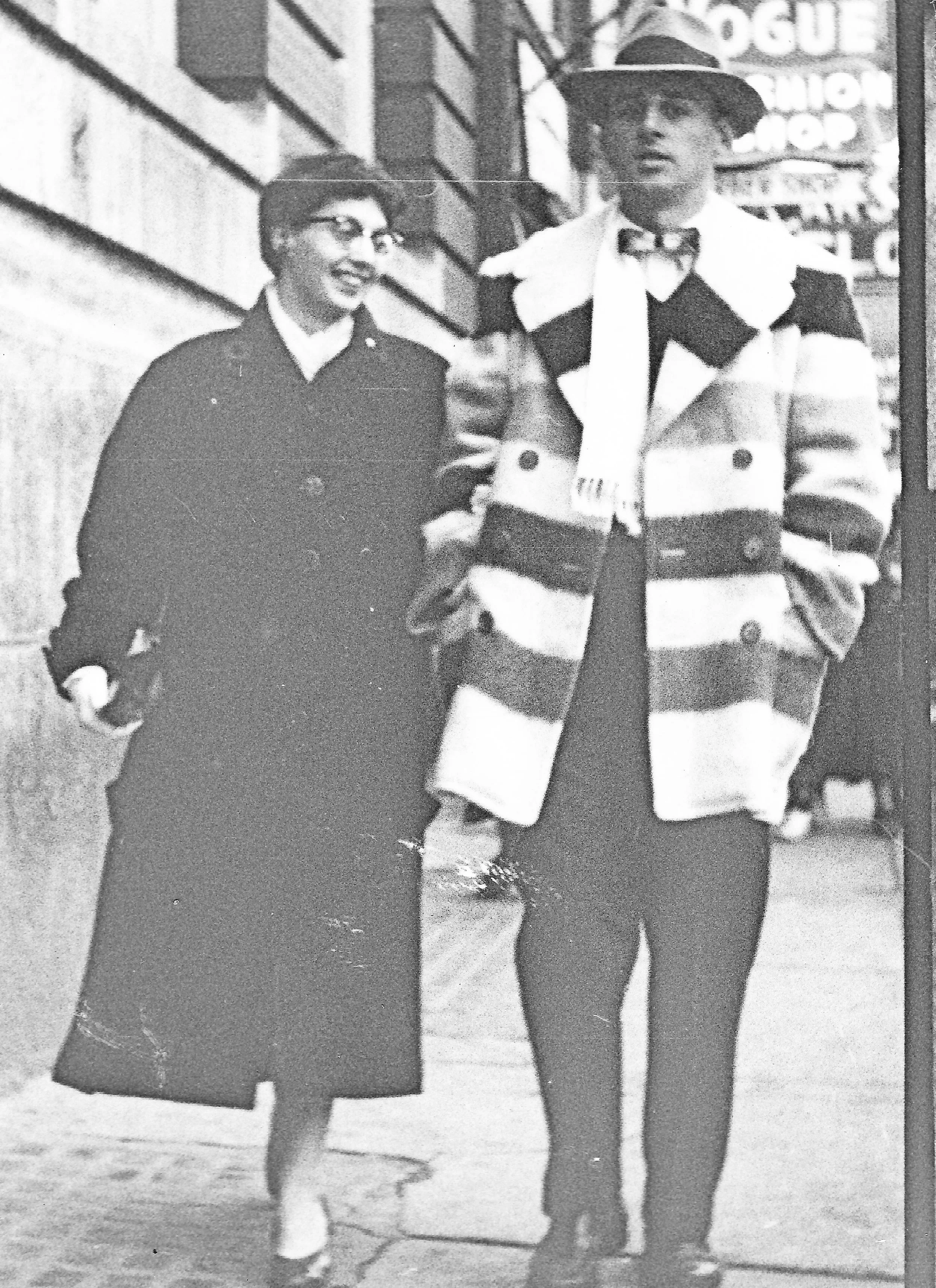 A smiling man and woman walk down the street. The woman has a long black coat and wears gloves. The man wears a snap-brim hat, a striped hudson's bay coat, and a silk scarf.