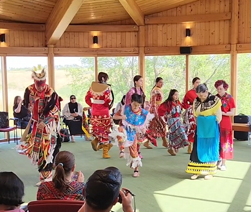 Several youth in Indigenous Regalia dance intertribal style. An elder with a shawl draped over her arm dances as well.