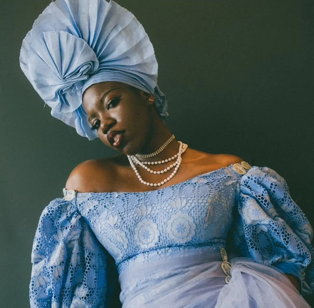 Peace Akintade-Oluwagbeye wearing a tall Gele and an elegant lace gown with puffed-sleeves, off-the-shoulder, both in matching ice-blue. She wears pearls around her neck.