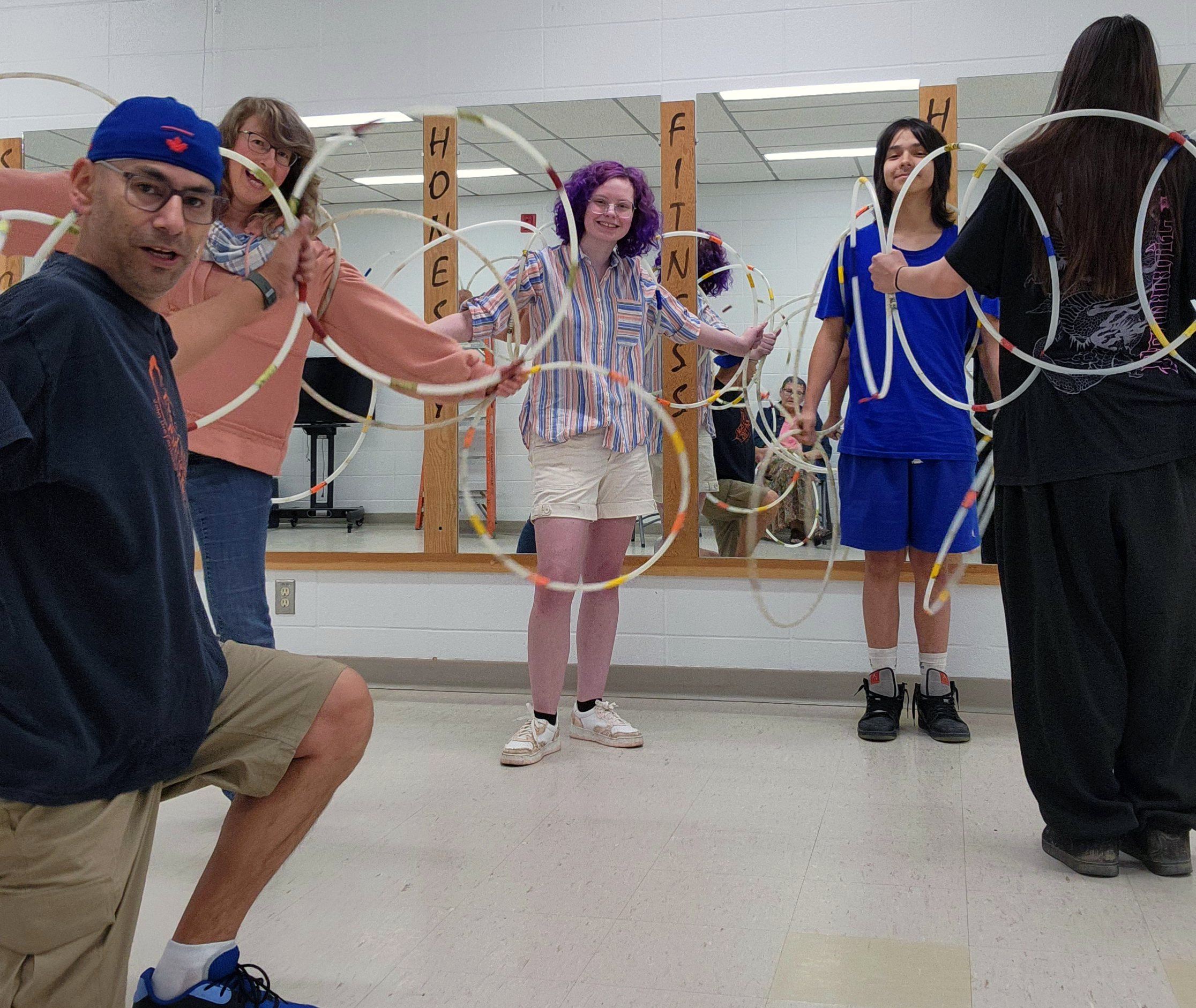 A man holds a camera for a selfie. He is kneeling in front of three youth and one adult woman. They are all holding hoops and smiling. One youth has their back to the camera, showing the pattern of hoops.