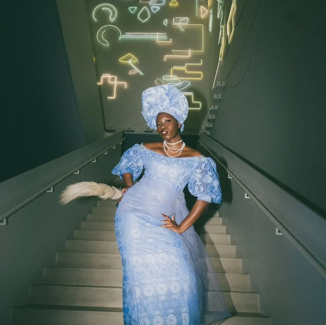 Peace Akintade-Oluwagbeye wearing a tall Gele and an elegant lace gown with puffed-sleeves, off-the-shoulder, both in matching ice-blue. She wears pearls around her neck. She is standing in a stairwell and cairries a feather fan.