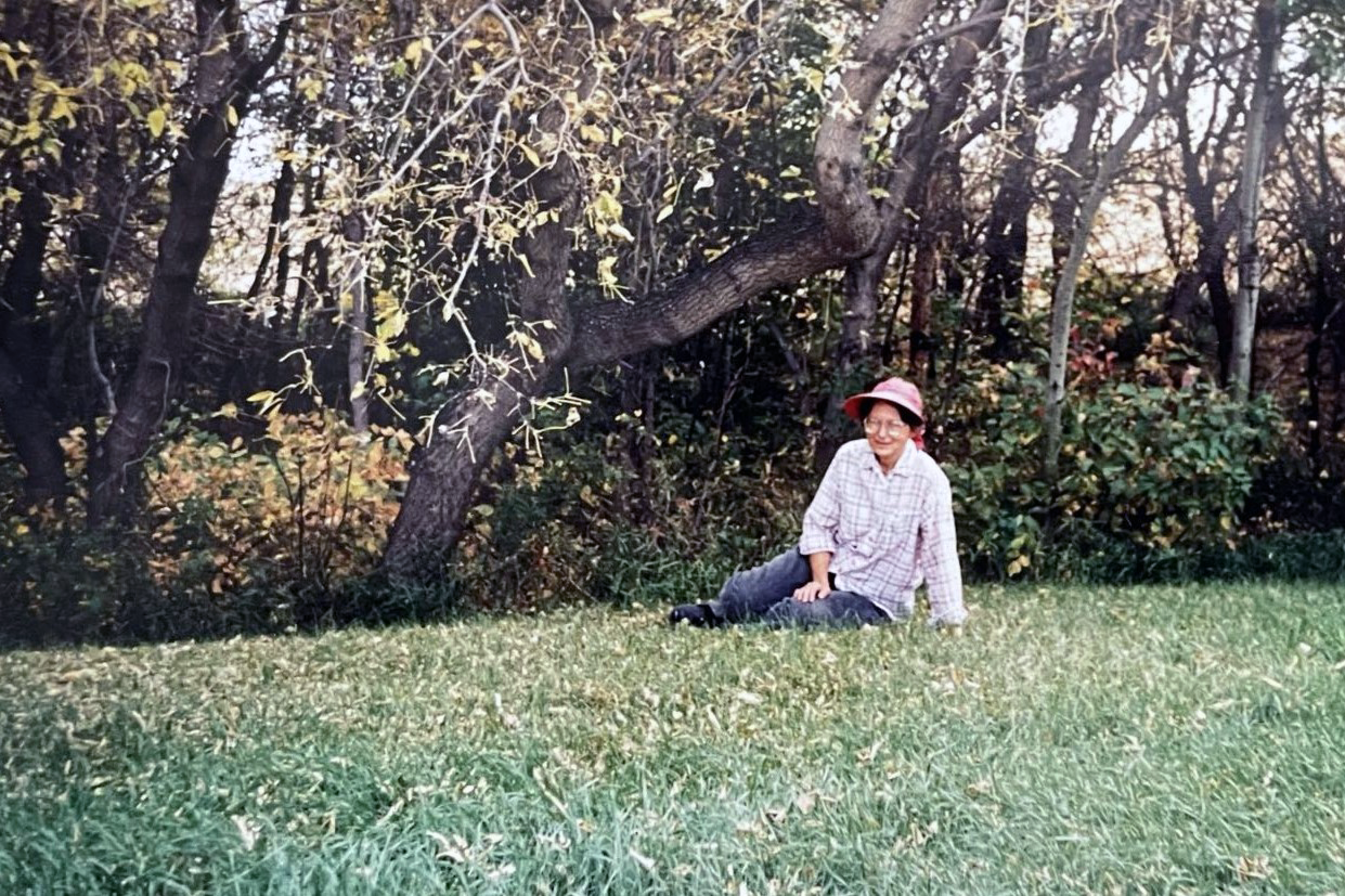 A woman in a plaid shirt, sun hat, and work pants sits on a green lawn with trees and brush in the background.