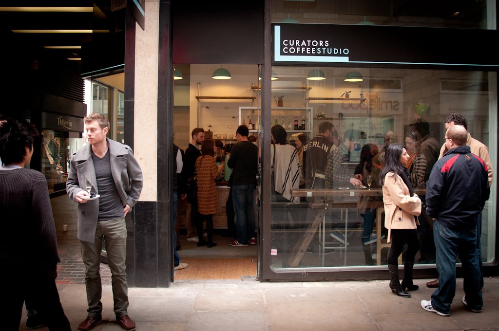 A group of people at a coffee shop, some people are inside ordering at the counter, and other people are standing outside drinking cups of coffee.