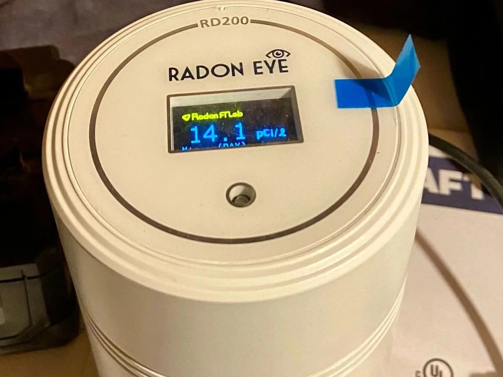 Close-up of a Radon Eye radon testing device with a digital screen and a blue sticker attached to the top right corner.