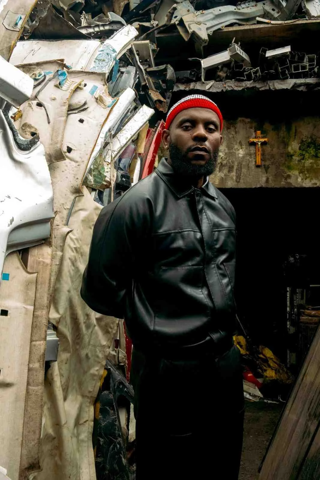 Odumodublvck wearing a black leather jacket and red beanie standing in front of a large pile of car wreckage.