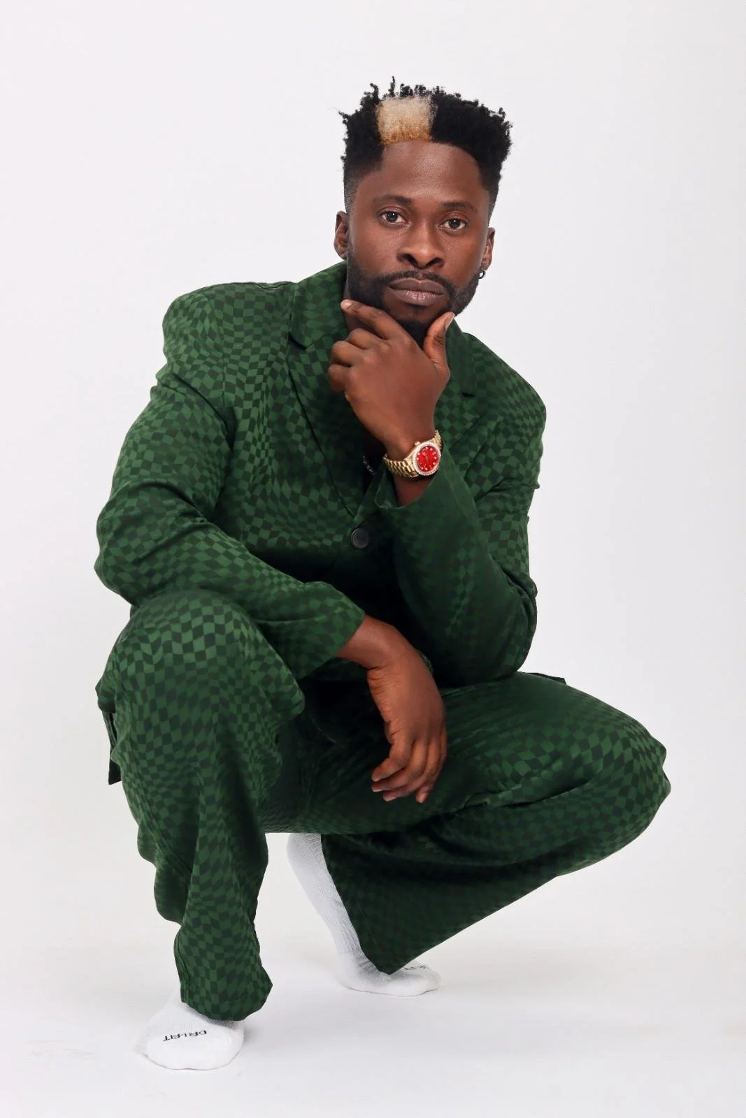 Dj Ecool flyer in a checkered green suit poses against a white background, squatting with one hand on his chin and the other on his knee, wearing a red watch and white socks.
