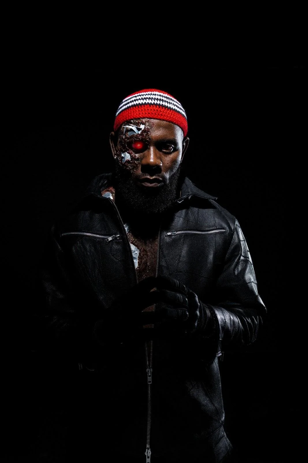 Odumodublvck in a black leather jacket and gloves wears a red, white, and black striped knit cap. He is positioned in front of a black background,  giving a cyberpunk or futuristic appearance.
