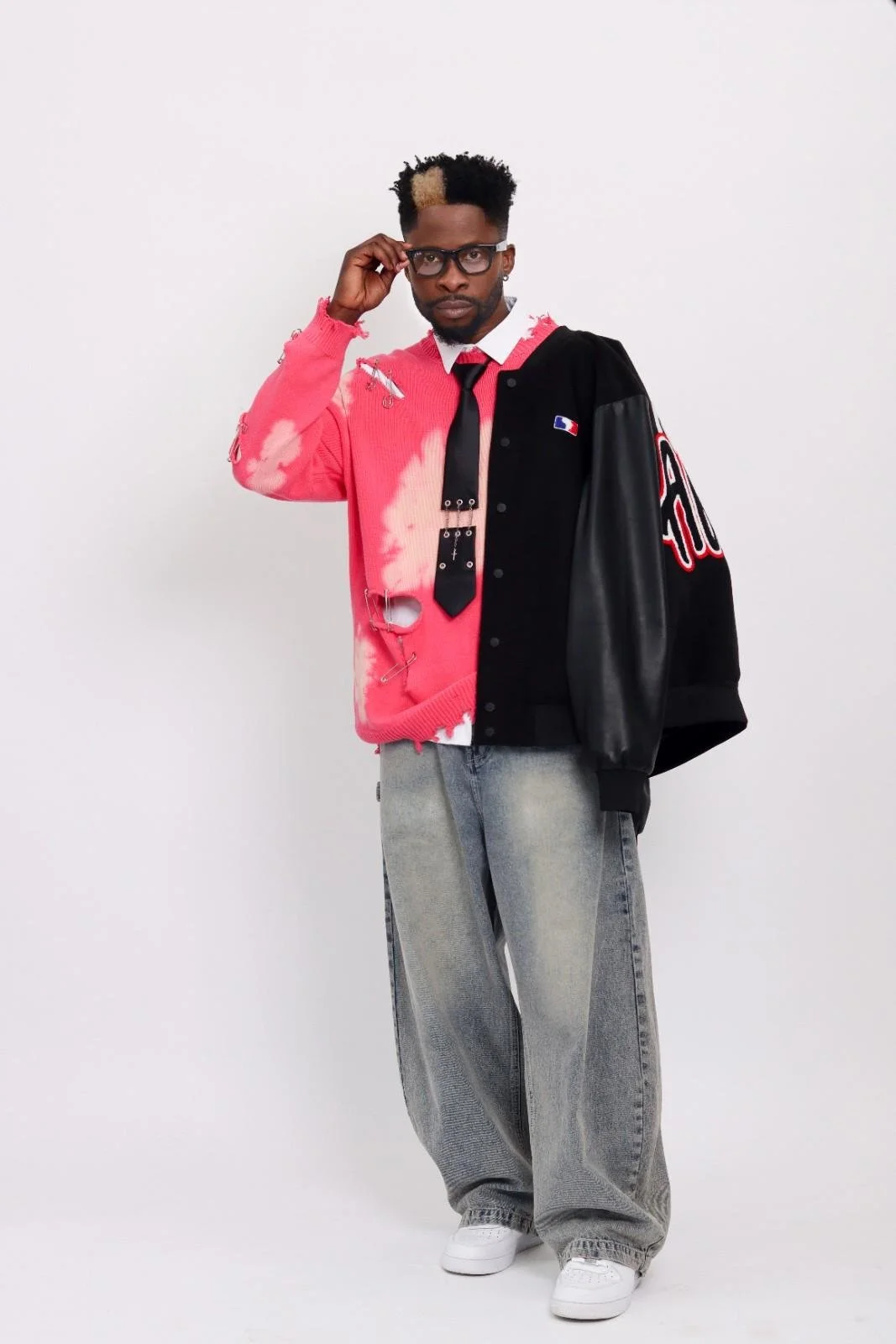Man wearing a distressed pink and black varsity jacket, grey baggy jeans, white sneakers, glasses, and a black tie, standing against a plain white background.