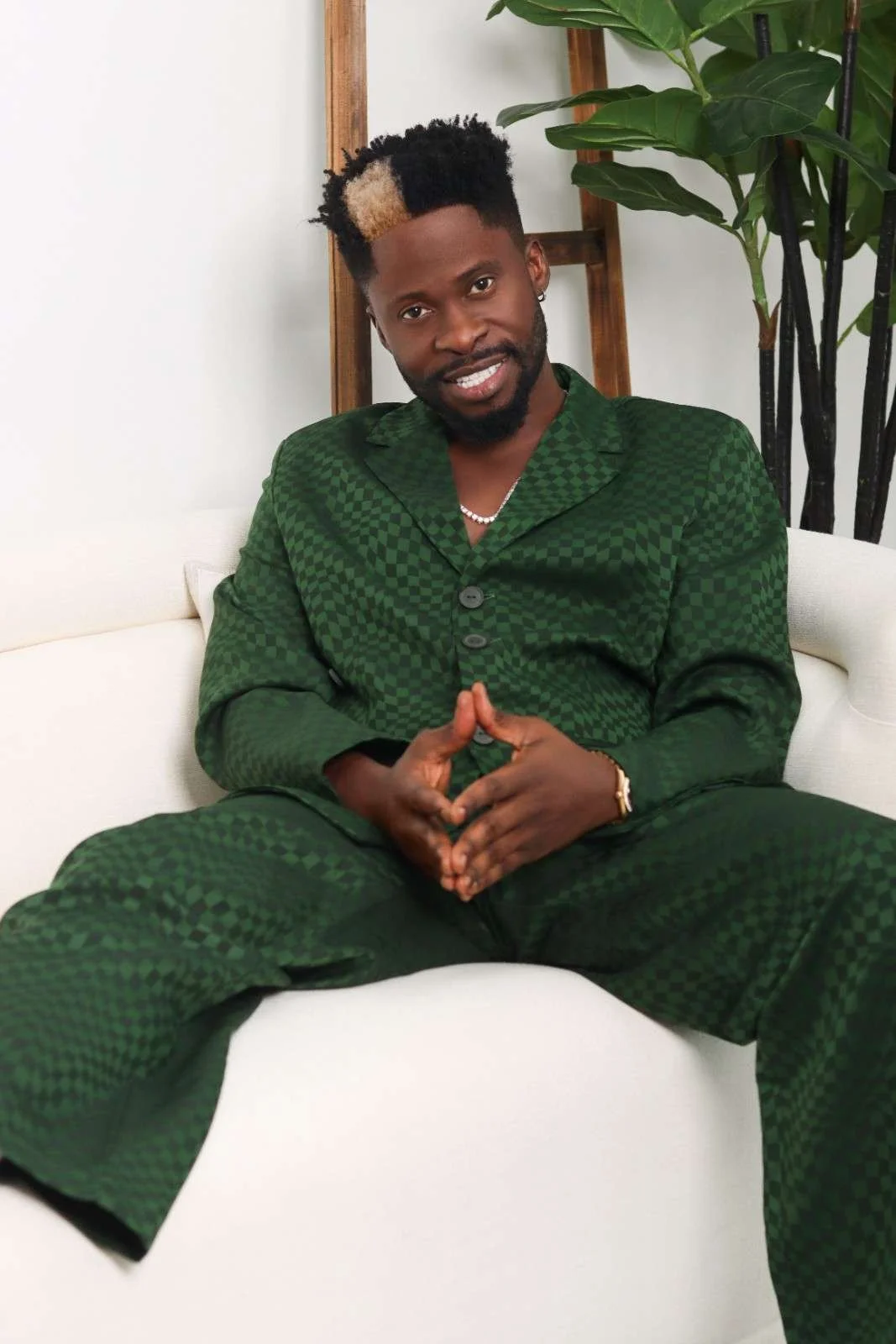Dj Ecool with short curly hair and a beard, wearing a dark green checkered suit, sits on a white couch with legs apart and hands clasped. He is smiling and looking at the camera, with a tall plant and a wooden frame in the background.