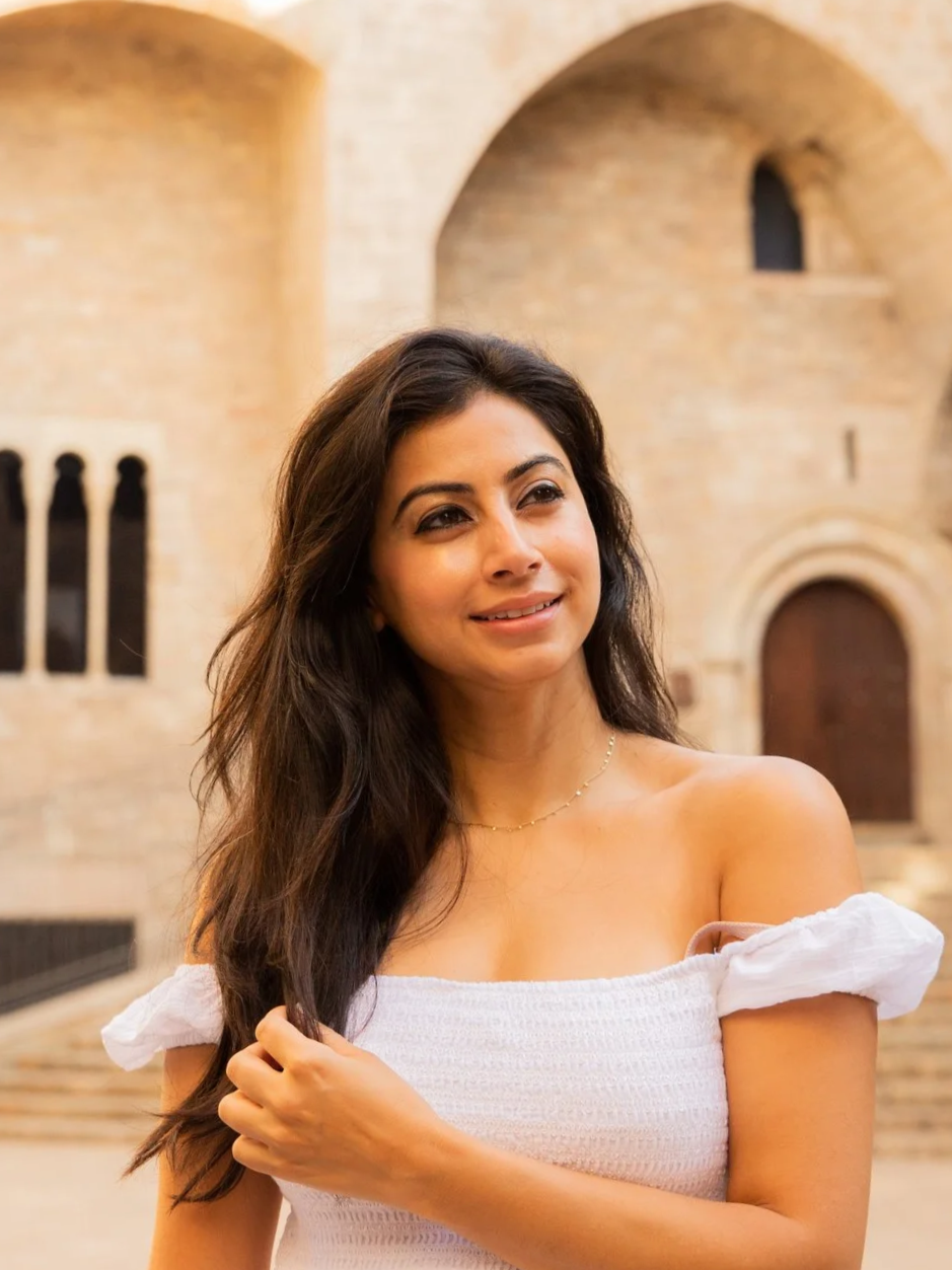 A woman with long dark hair wearing a white off-shoulder top standing outdoors with a historic stone building in the background.
