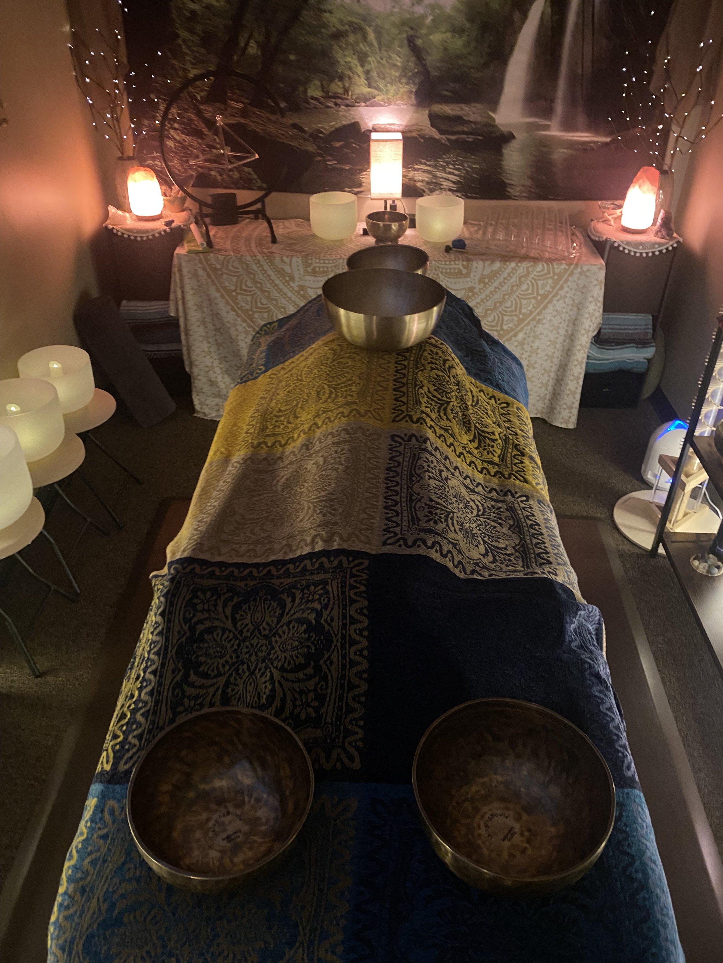 A relaxing meditation room with a decorated massage bed, Himalayan salt lamps, crystal bowls, and ambient lighting. A forest waterfall mural covers the wall.