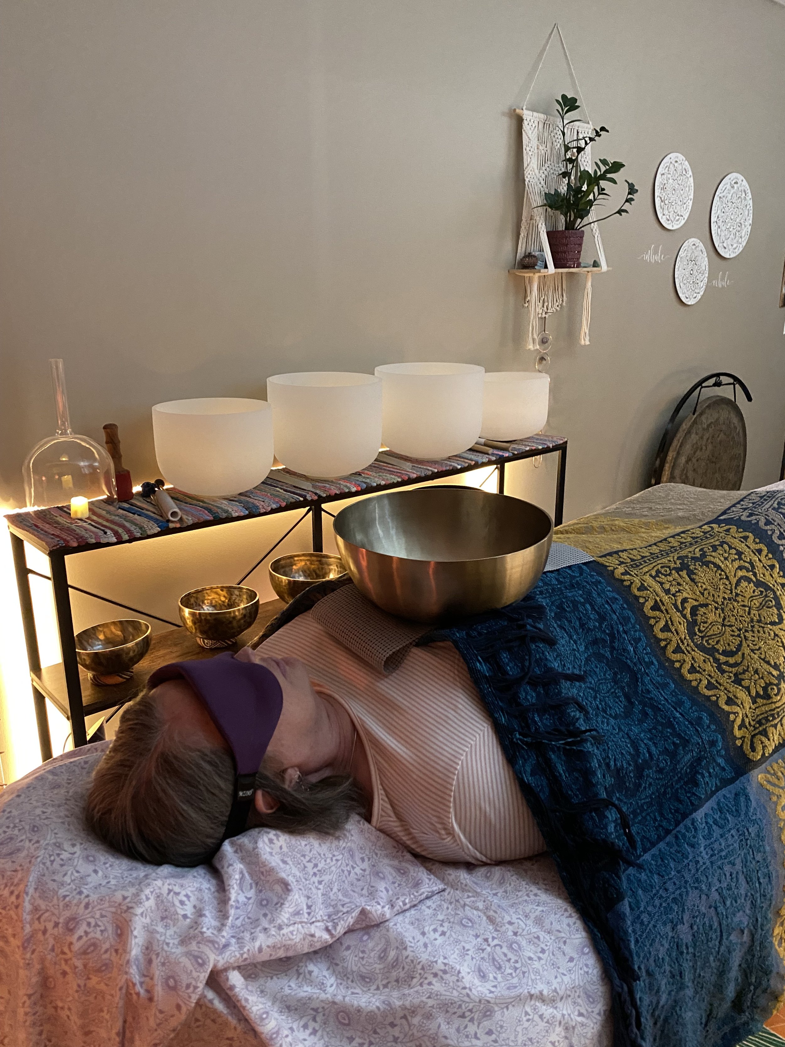 A person lying on a treatment table covered with a blanket, wearing an eye mask, with a Tibetan singing bowl on their chest. In the background, there are more singing bowls on a shelf, along with decorative items on the wall.
