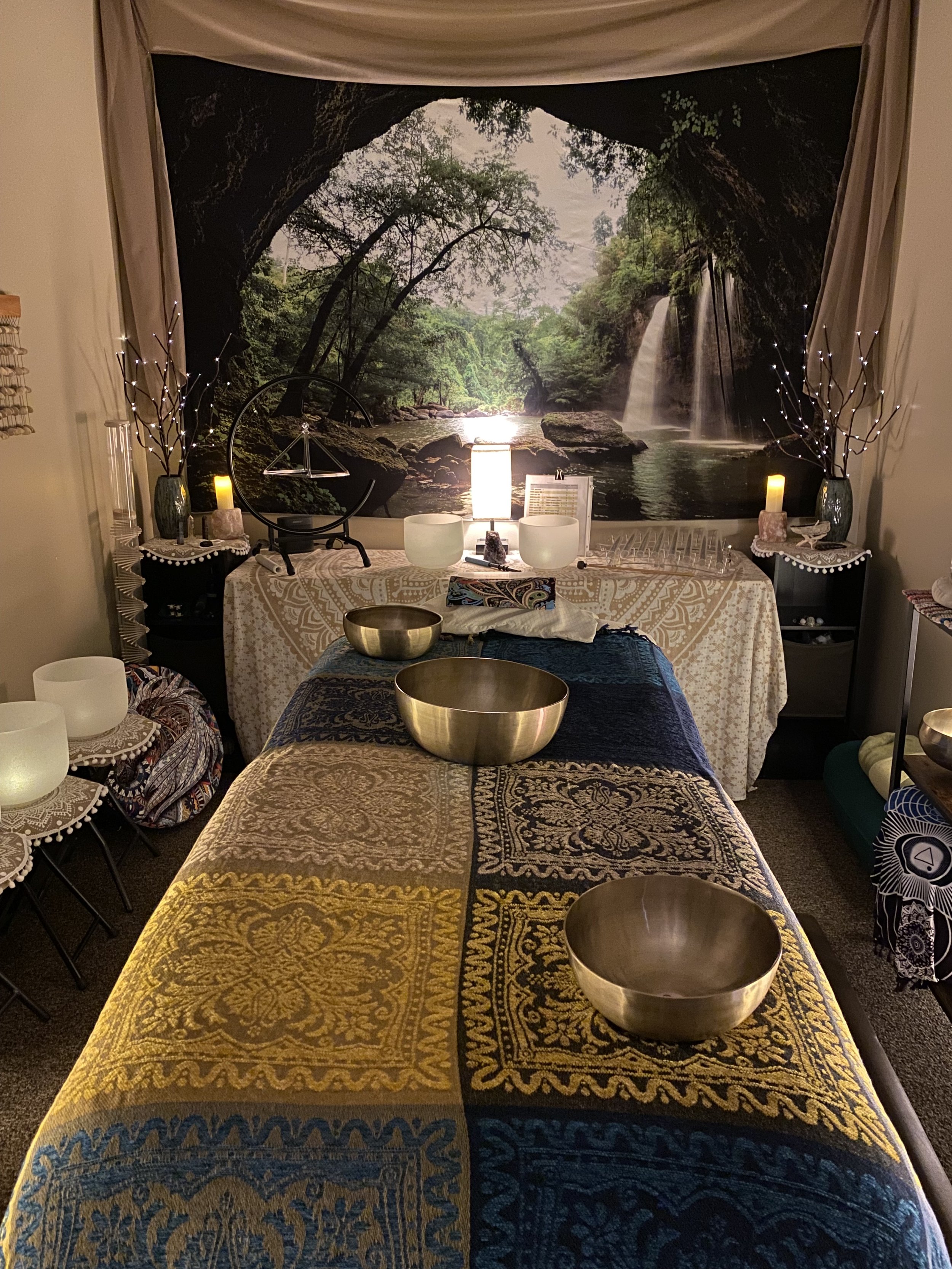 Interior of a tranquil room with a massage table covered in a patterned blanket, adorned with singing bowls. A nature-themed tapestry with a waterfall scene hangs on the wall. The room is softly lit with candles and decorative lights, creating a calm