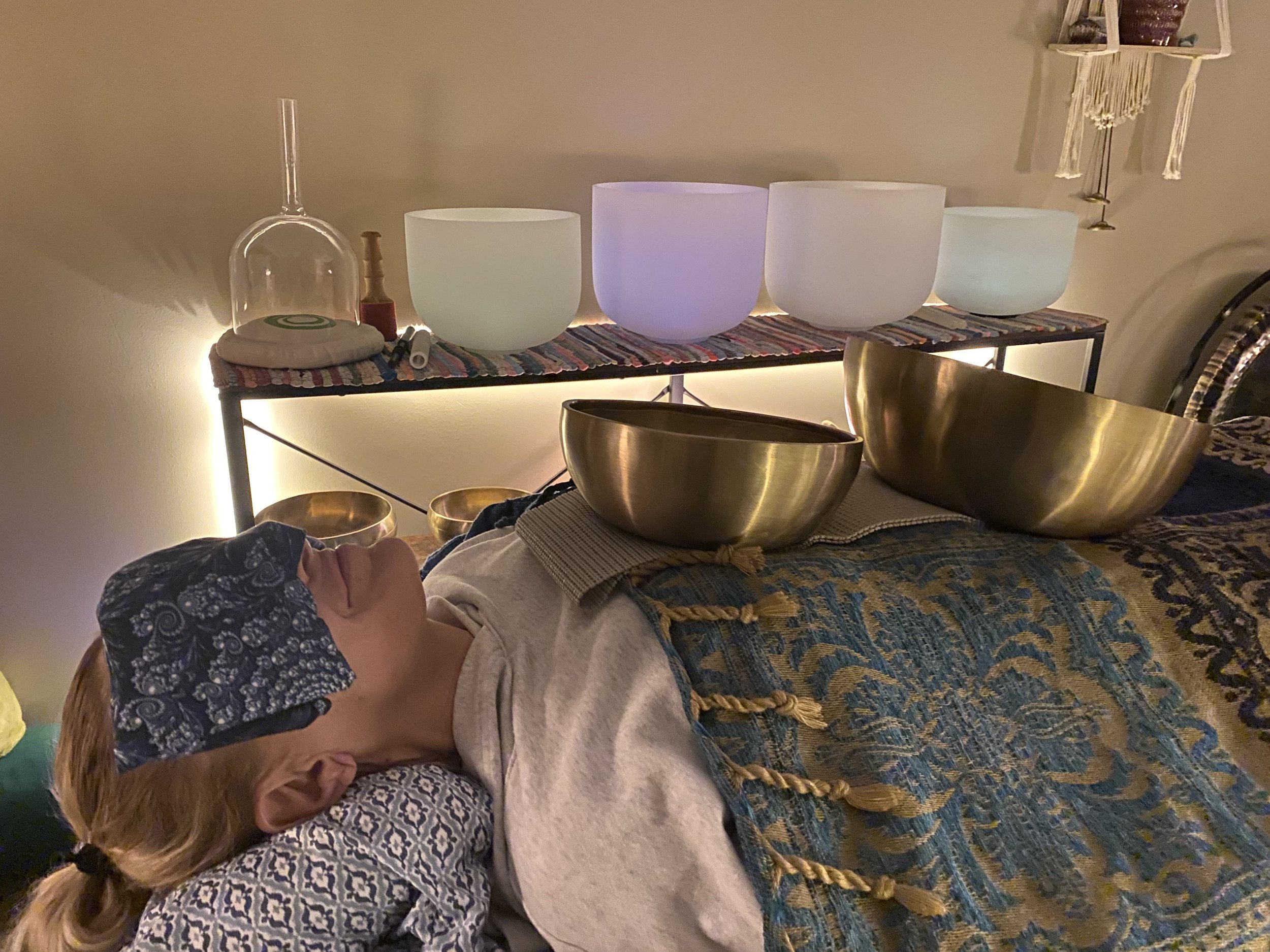 Person lying down in a cozy room with Tibetan singing bowls on their body, wearing an eye mask, surrounded by crystal singing bowls and ambient lighting, suggesting a sound healing session.