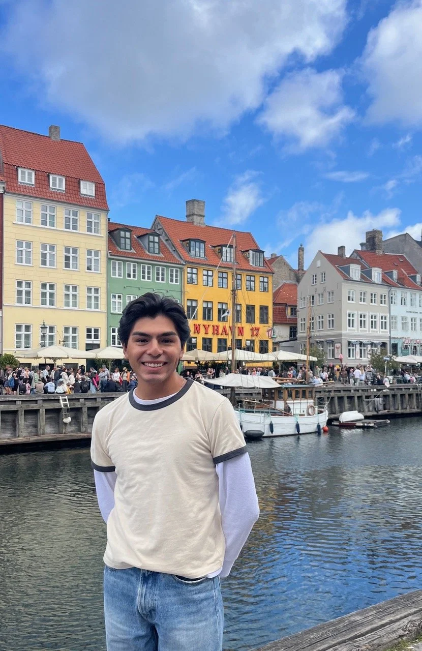 A young man with dark hair smiling at the camera standing by a canal with colorful buildings and a crowd of people in the background.