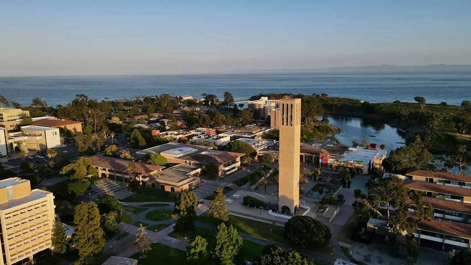 Aerial view of a UCSB campus with a tall clock tower, buildings, trees, a river, and the ocean in the background during sunset.