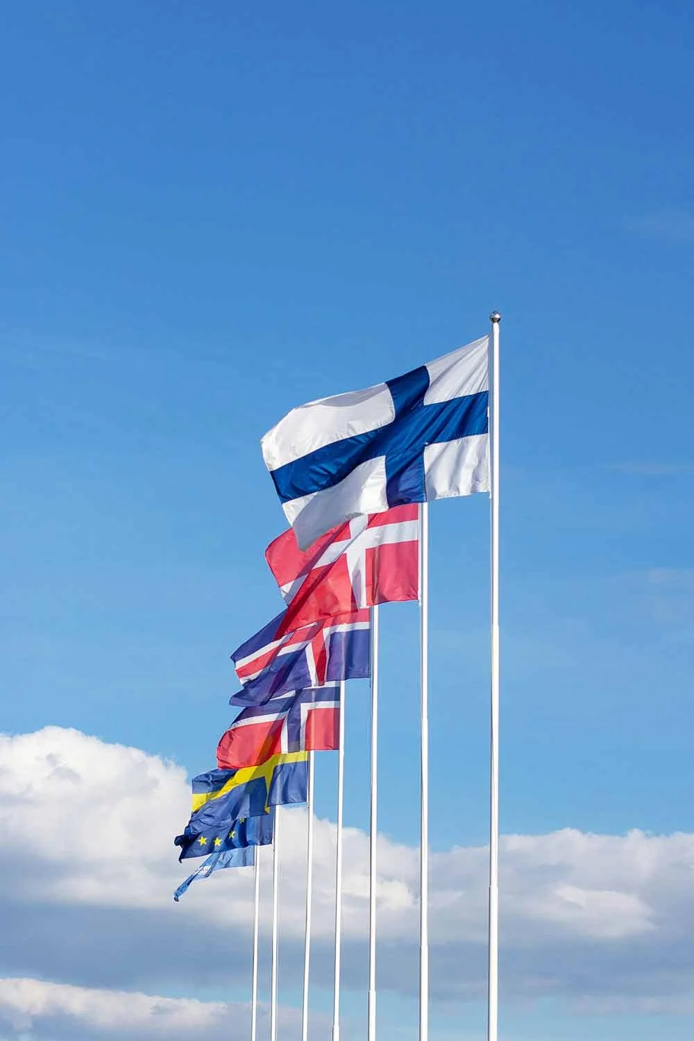 Six Scandinavian flags flying in the wind against a blue sky with some clouds.