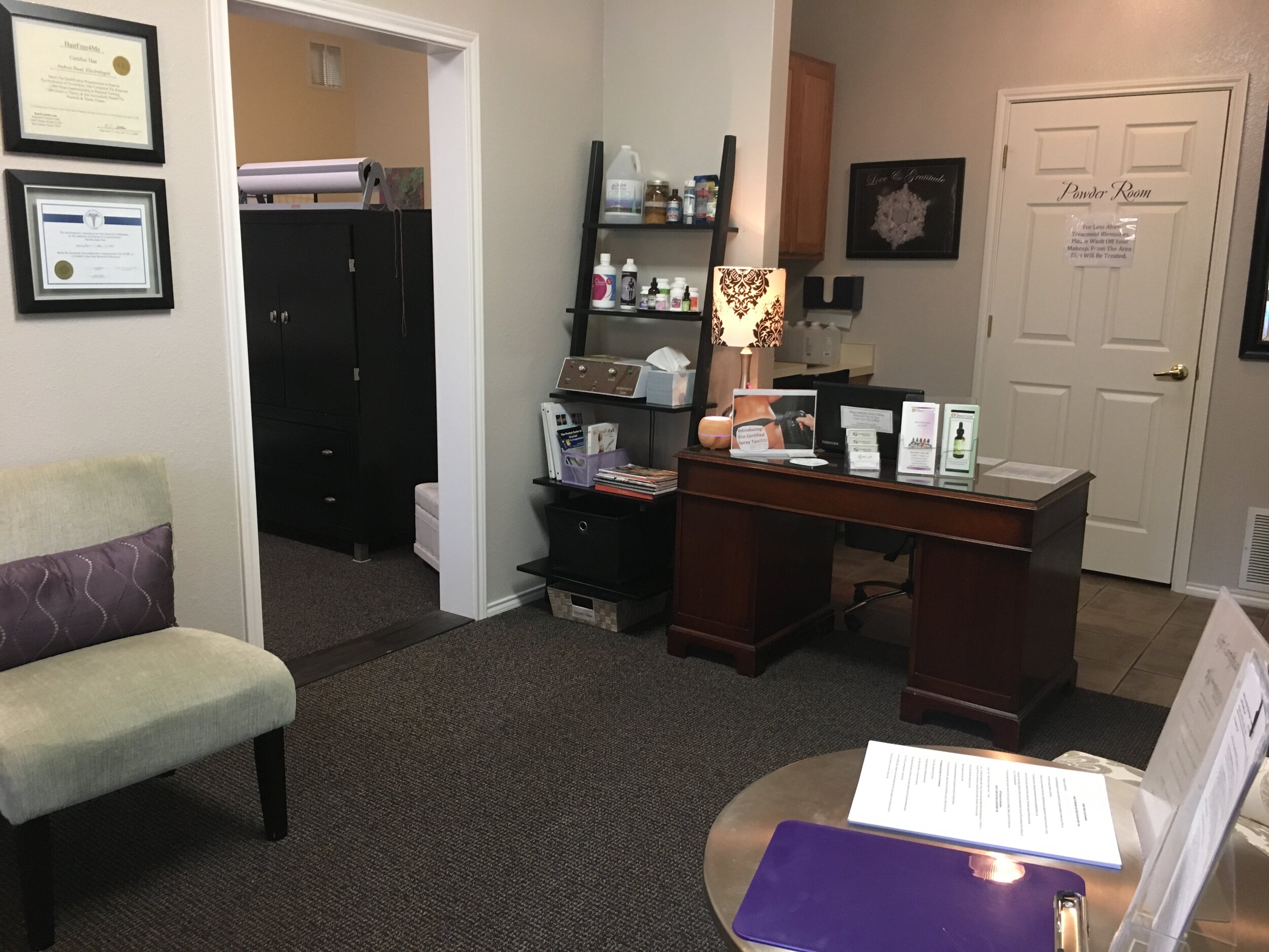 Interior of a clinical office with a wooden desk, a lamp, a bookshelf with medical supplies, framed certificates on the wall, and a doorway labeled 'Powder Room'.