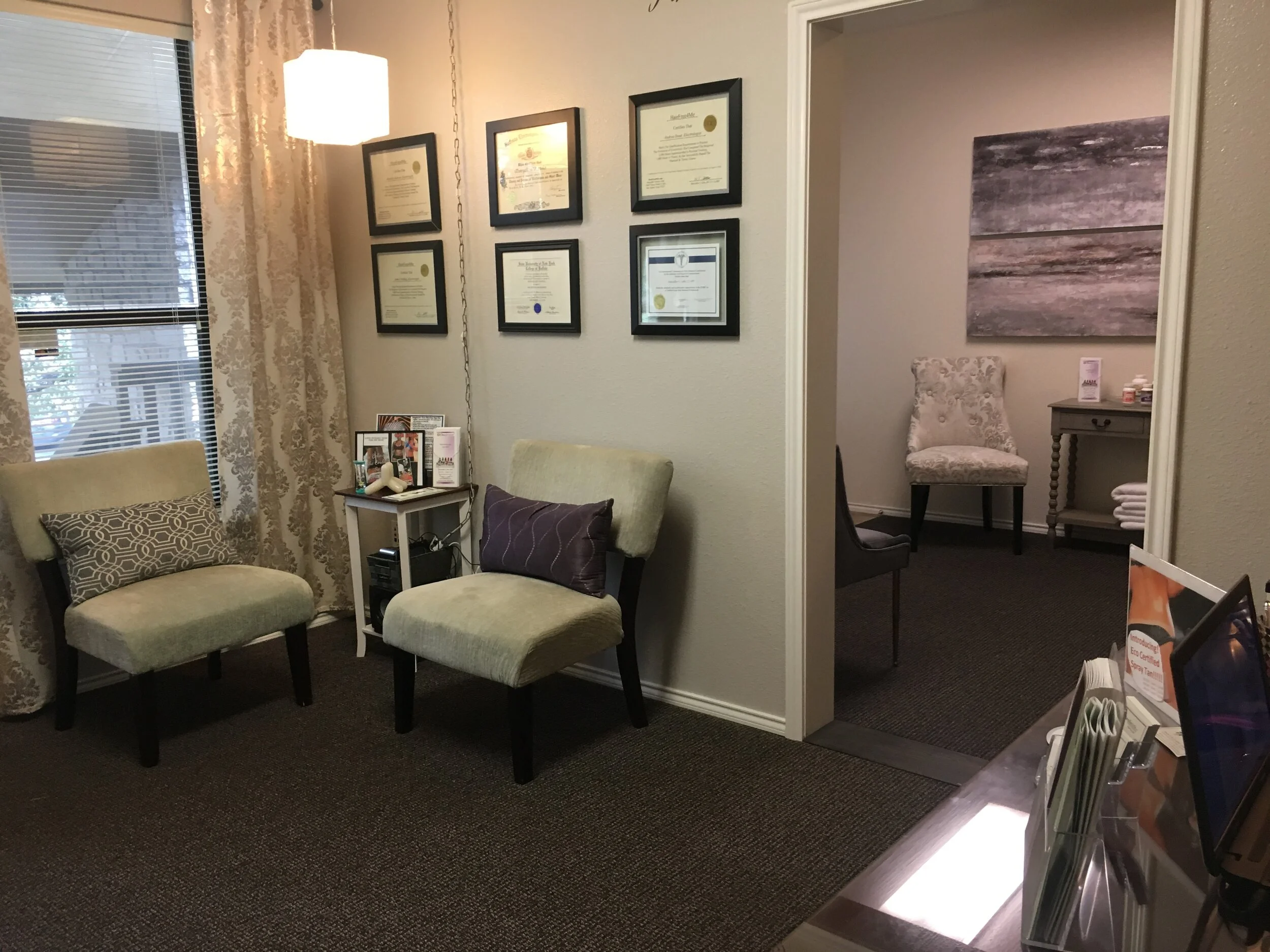 Waiting room with two chairs, framed certificates on the wall, window with blinds and curtains, side table with magazines and a small mixer, small room visible through doorway with a chair, artwork, and a table with folded towels and bottles.