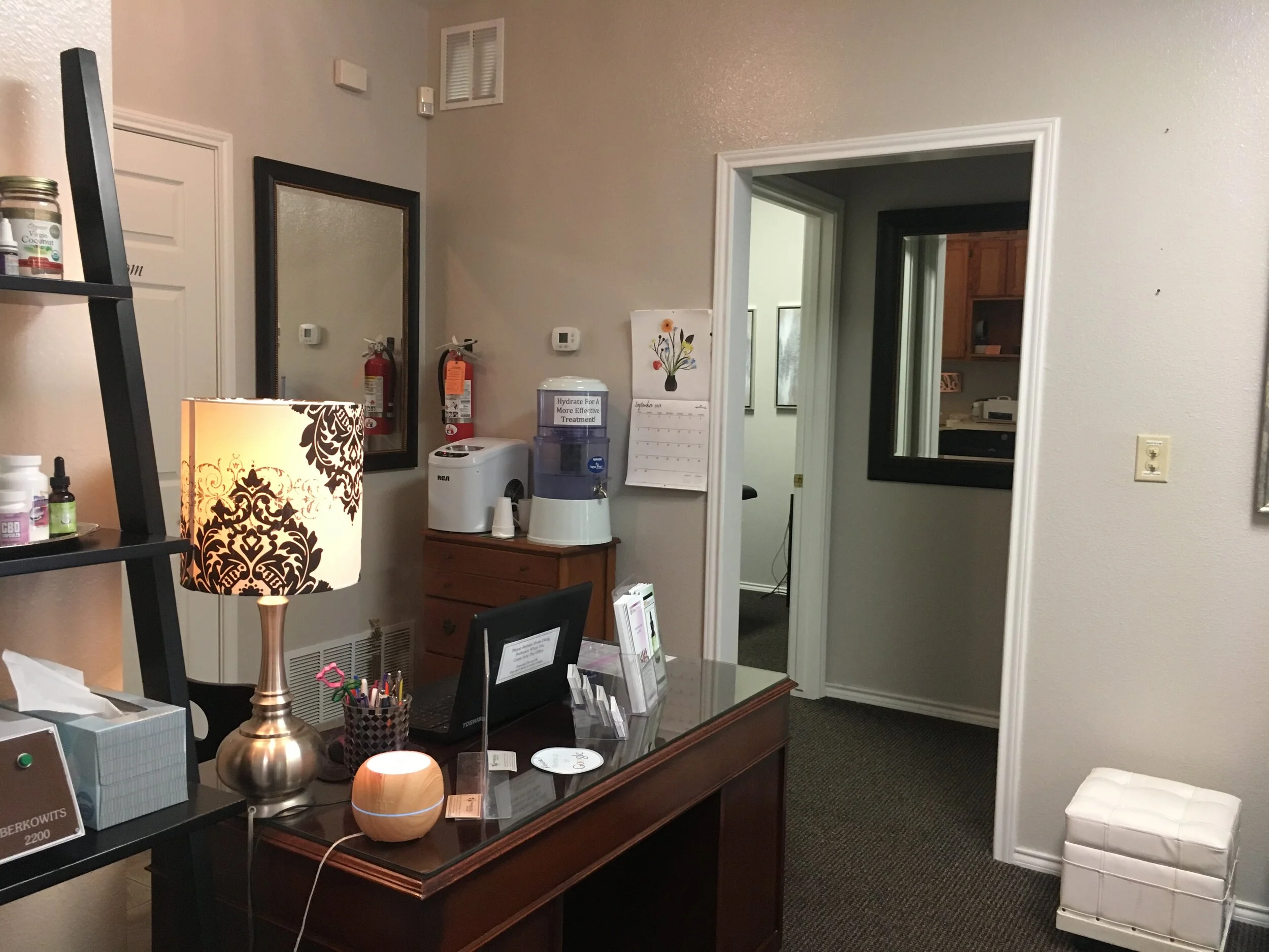 Office reception area with a dark wooden desk, a decorative table lamp, a small diffuser, and various office supplies, with a doorway leading to a kitchen area in the background.