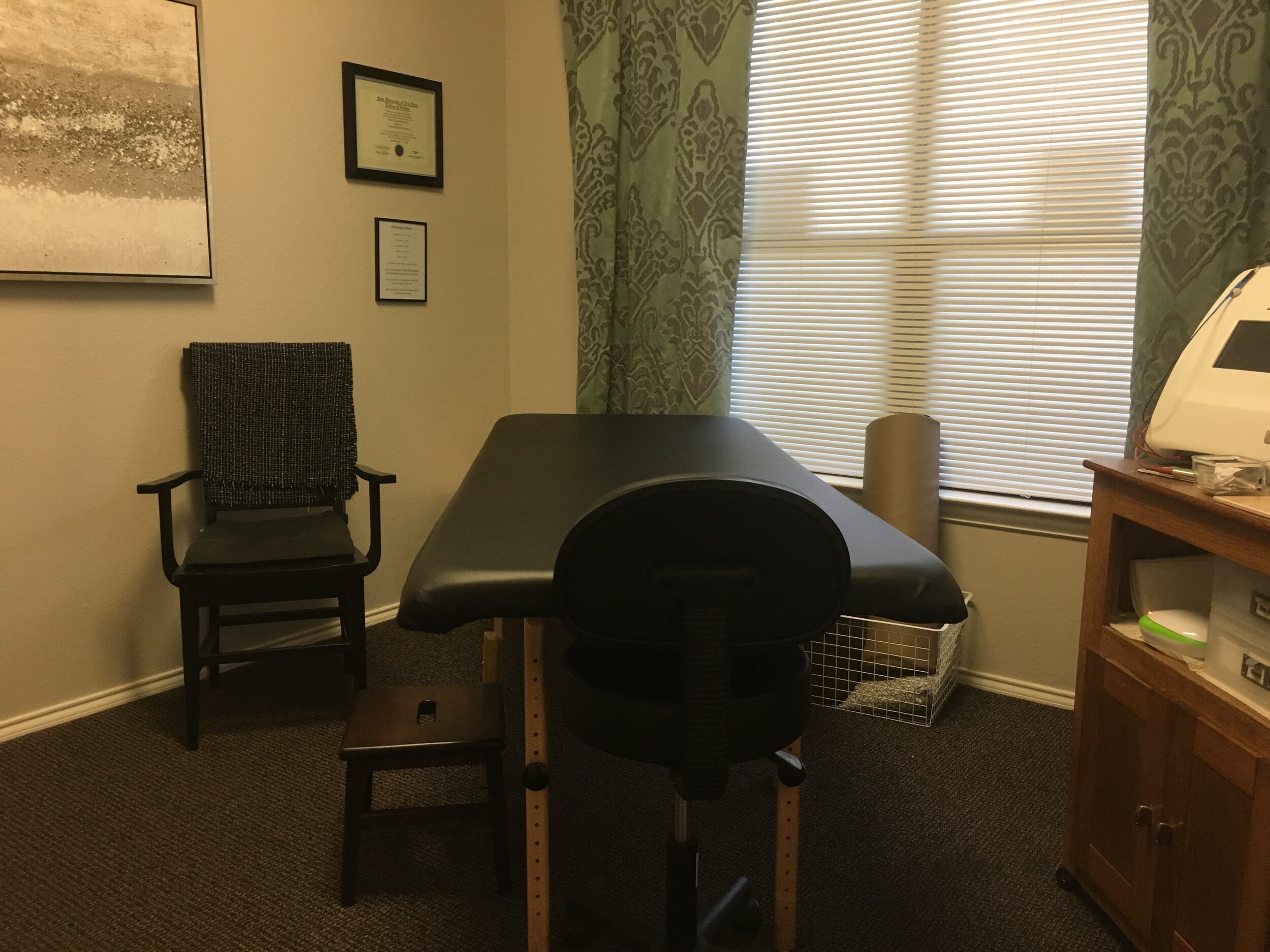 Massage or therapy room with a massage table in the center, a wooden cabinet on the right, a black chair and small bench on the left, framed certificates on the wall, window with blinds and curtains, and a medical device on the cabinet.