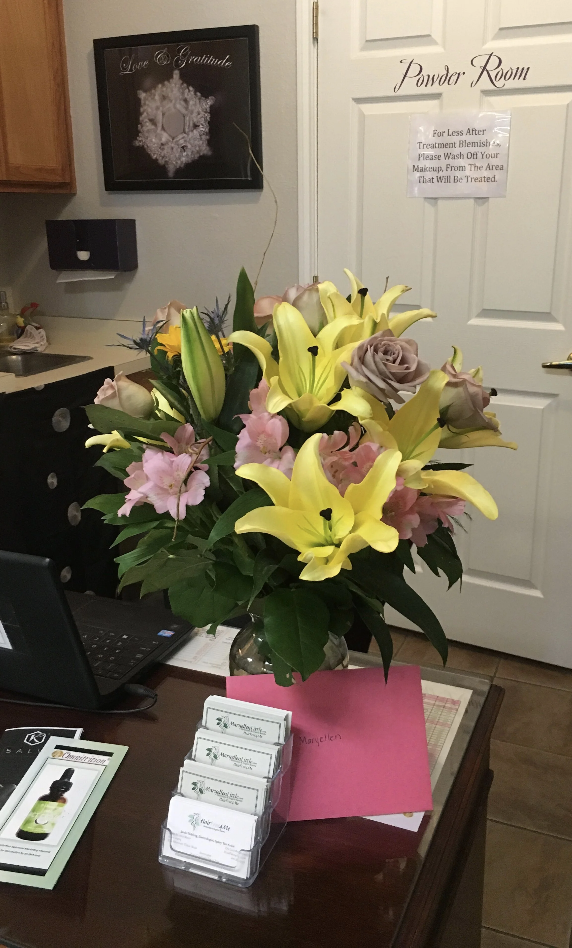 A large bouquet of yellow lilies, pink alstroemeria, peach roses, and purple roses in a clear glass vase on a wooden table. In the background, there is a sign on a white door that reads 'Powder Room' and a handwritten note below it. Various business 