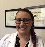 A woman with glasses and a braid smiling, wearing a white coat, standing in front of framed diplomas on a wall.