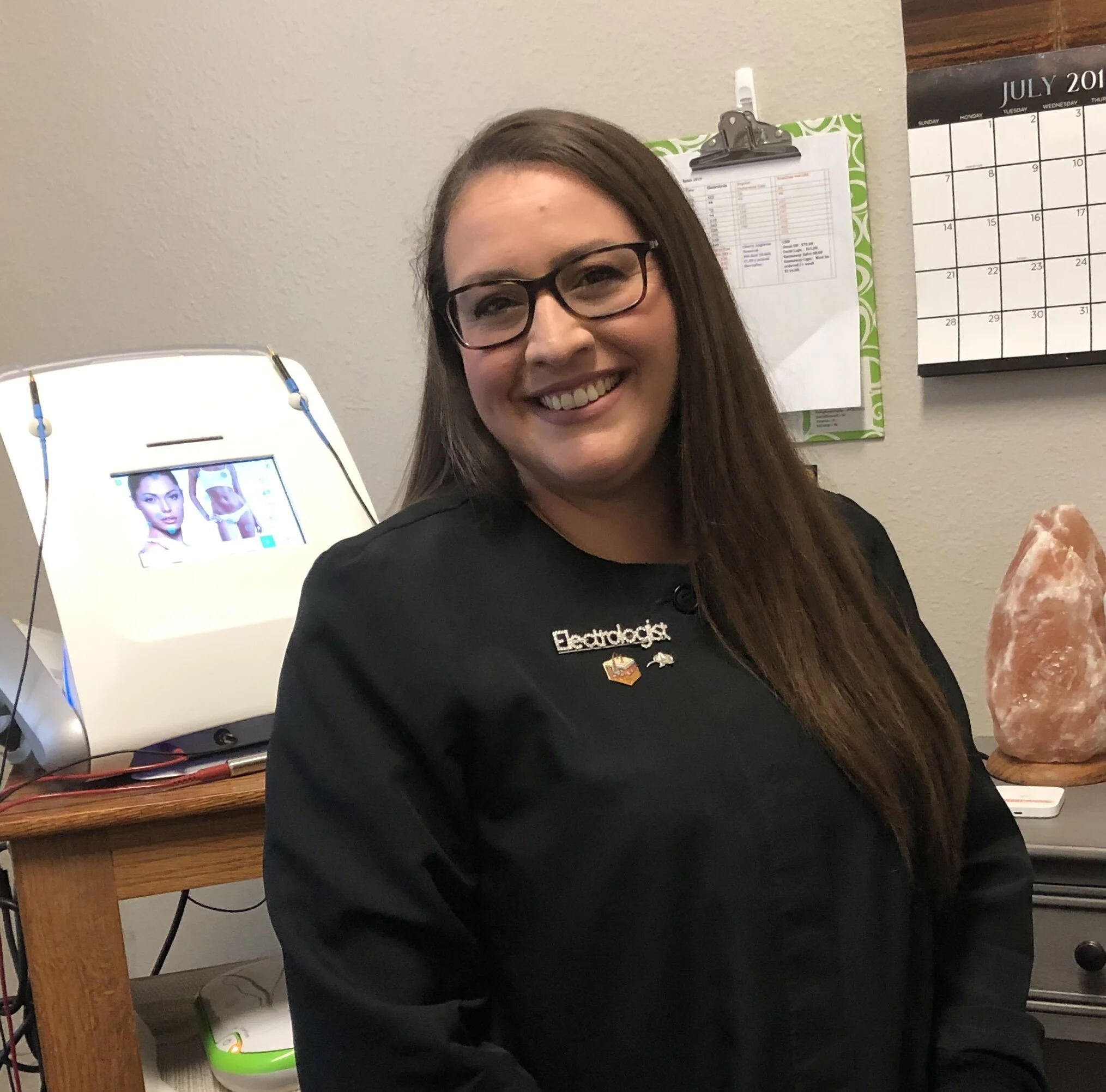 A smiling woman with long brown hair, black glasses, and a black clinical jacket with a name tag that reads 'Electrologist'. She is sitting in a room with a wall calendar displaying July 2019 and a green clipboard. Behind her is a salt lamp on a desk