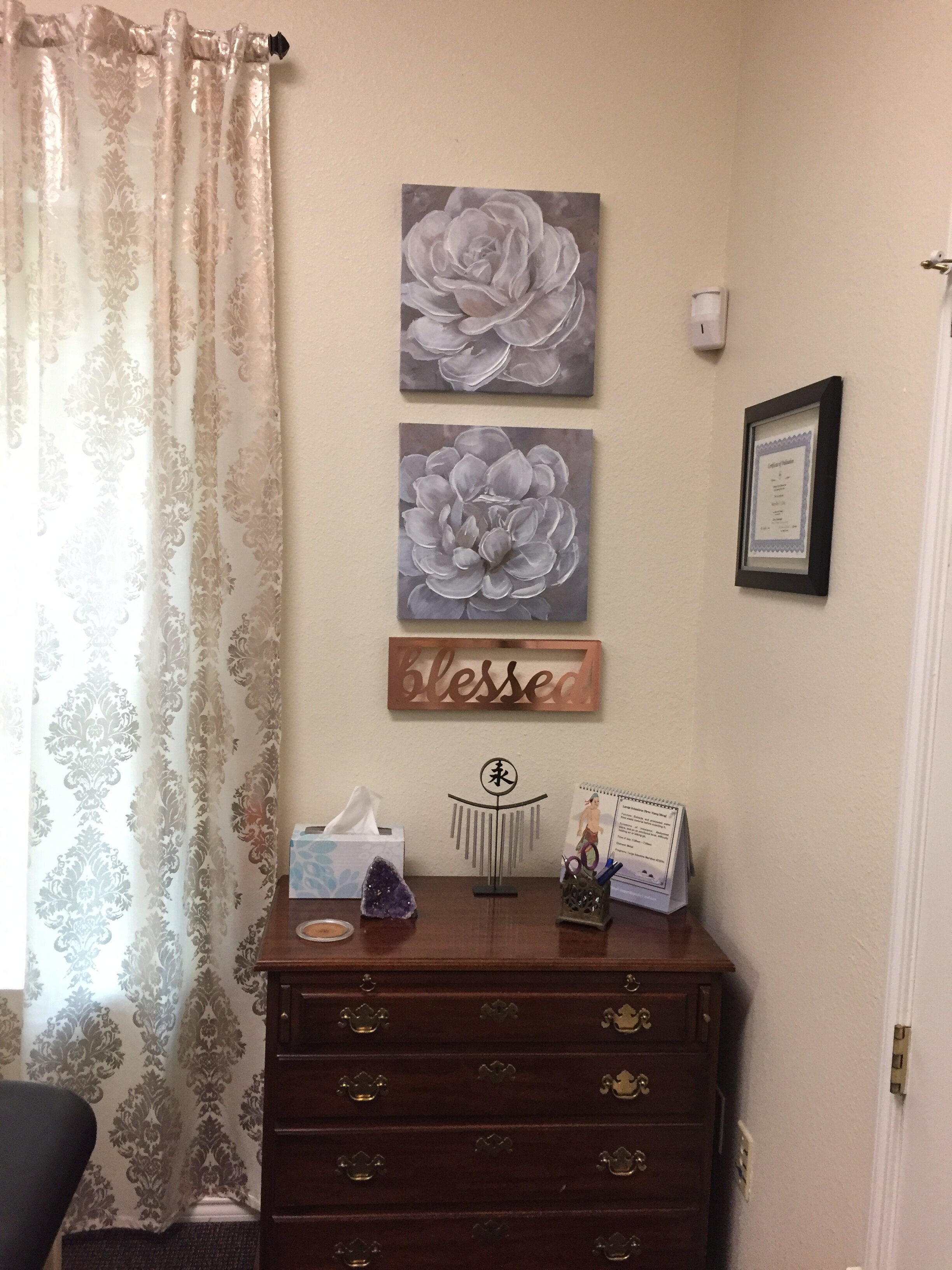 A wooden dresser with four drawers, on which sits a box of tissues, a purple crystal, a decorative metal piece, and a framed sign. Above the dresser are three square floral paintings in shades of white and gray, and a wooden sign that reads 'blessed'