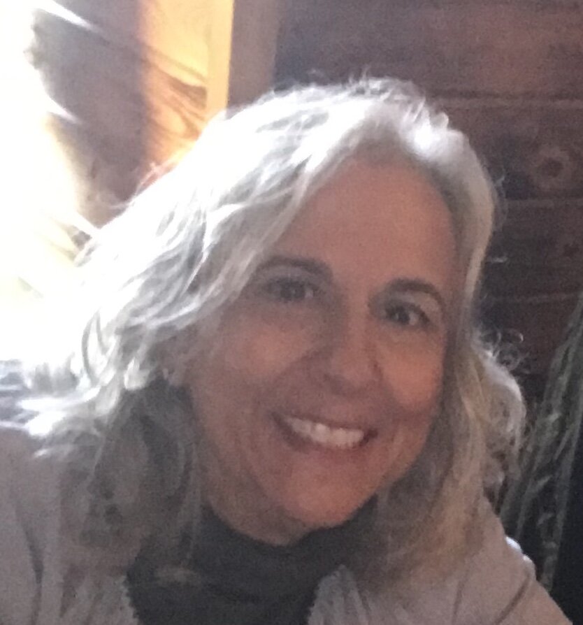 A woman with shoulder-length blonde hair smiling at the camera inside a room with wooden walls.