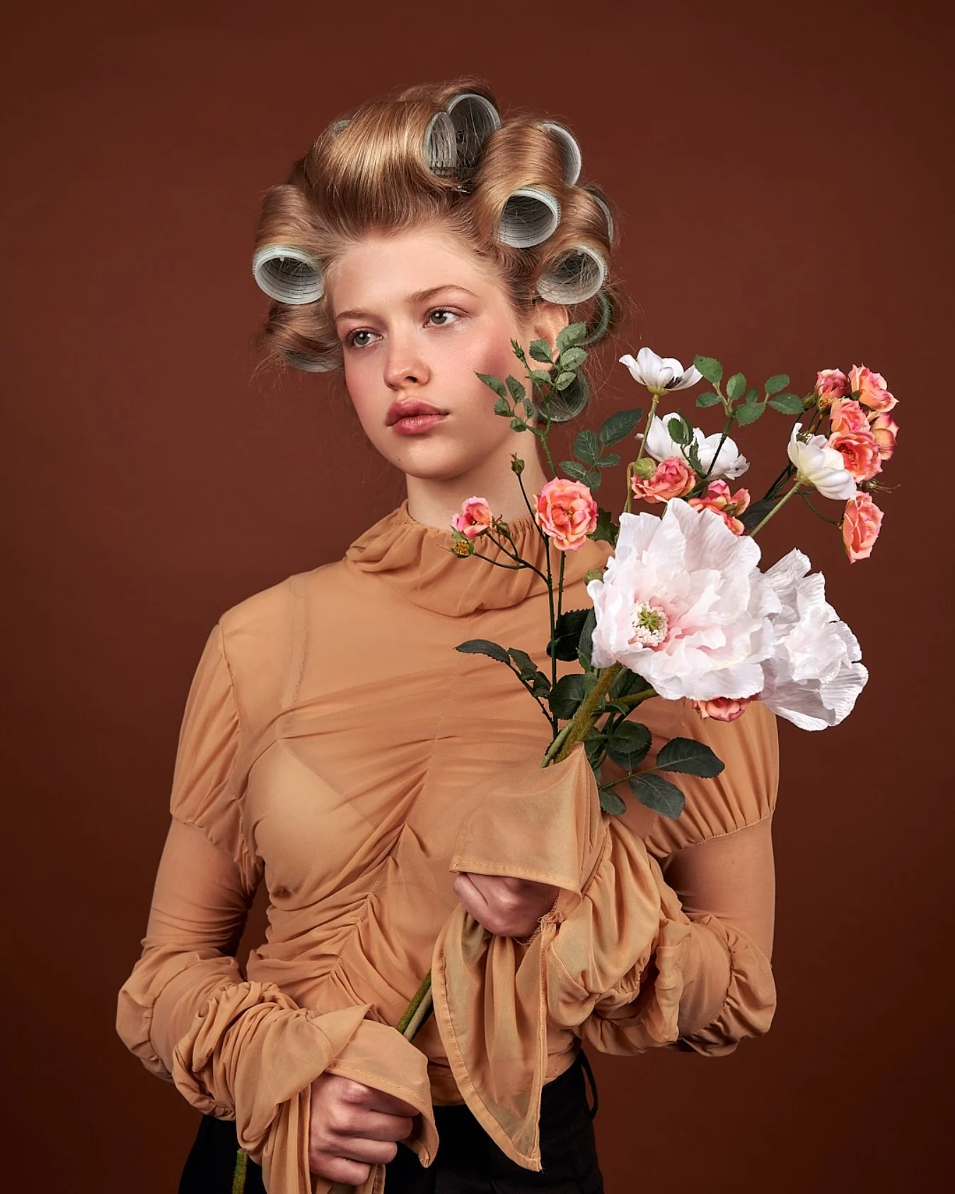 Young woman with rolled hair in curlers, wearing a beige sheer blouse with gathered fabric, holding pink and white flowers.