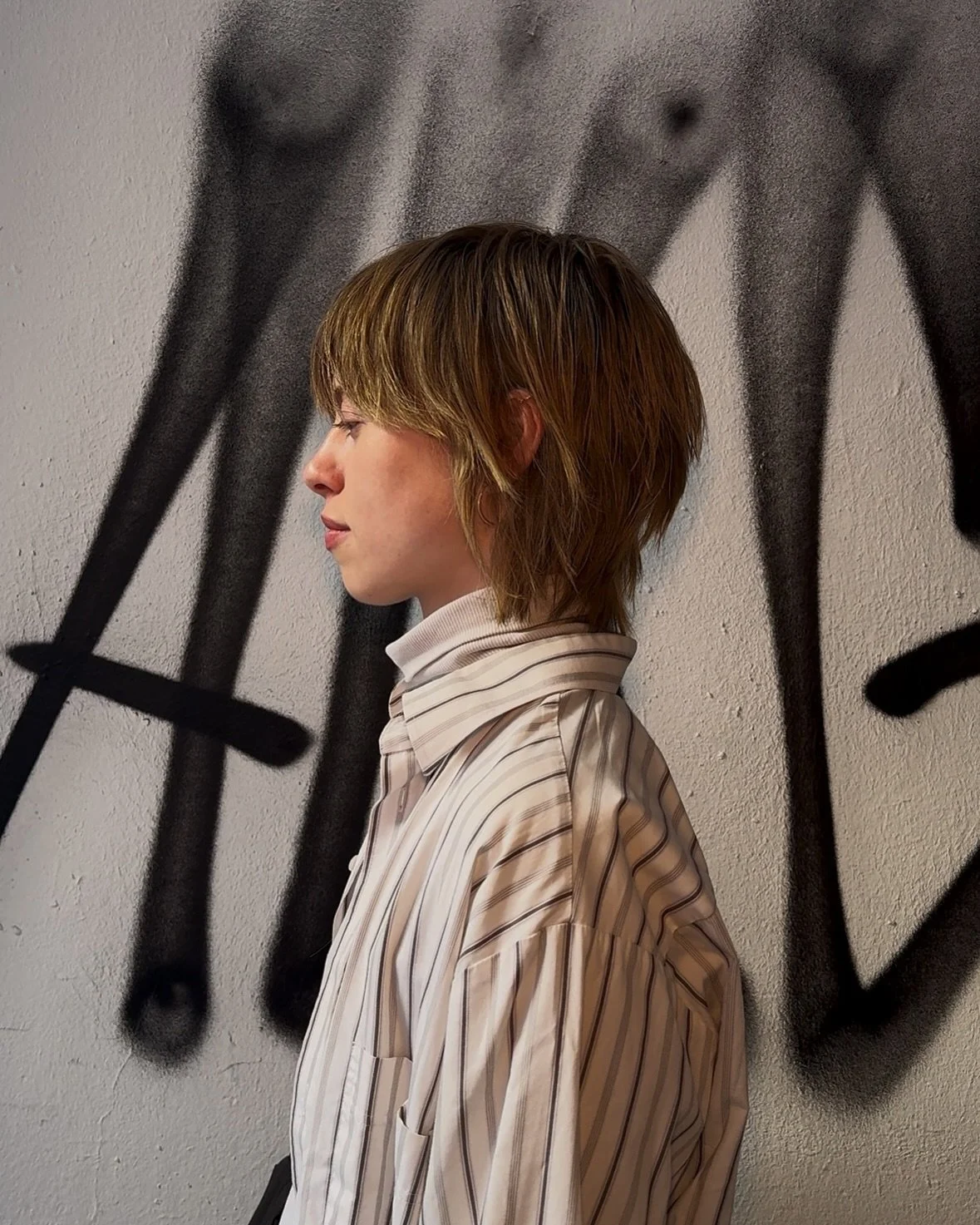 Side profile of a person with short, layered, dark blonde hair against a wall with black graffiti art on it.