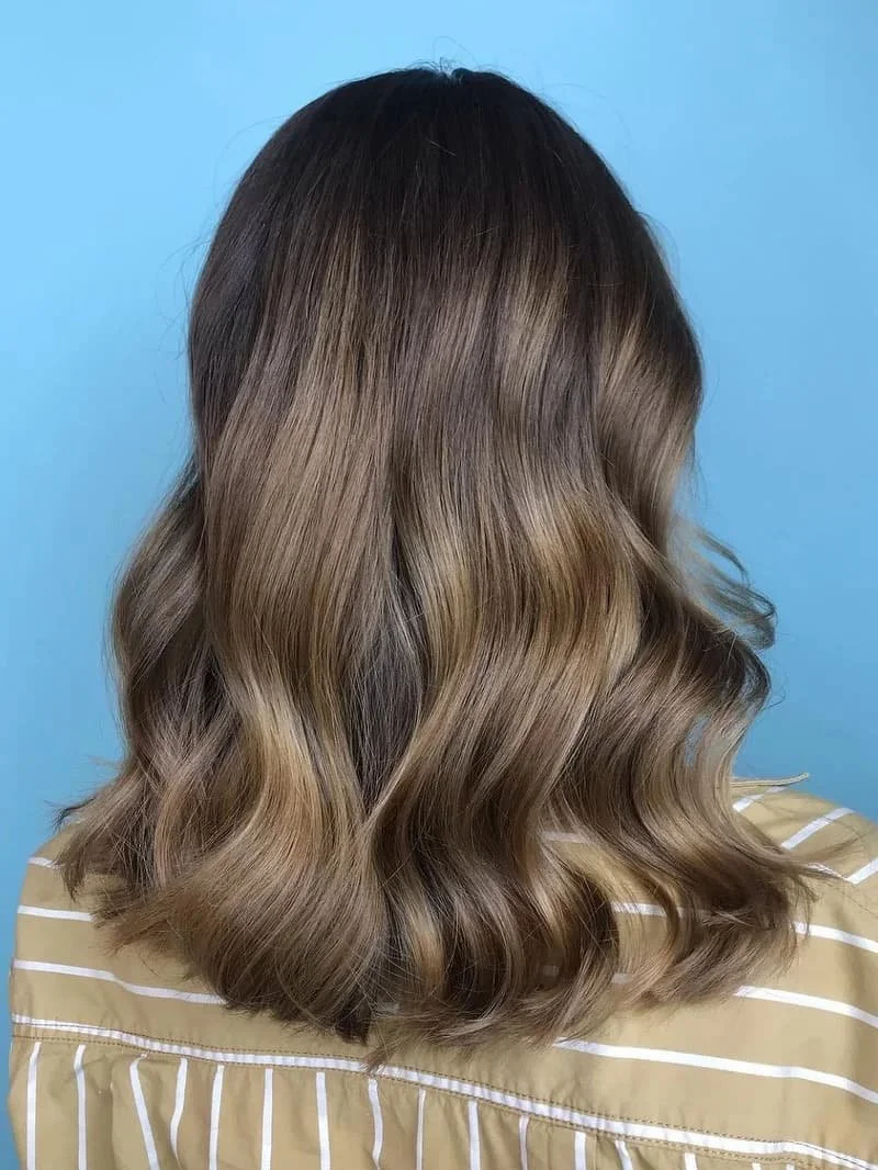 Back of a woman's head with shoulder-length wavy hair in shades of light brown and blonde, wearing a yellow and white striped shirt against a blue background.
