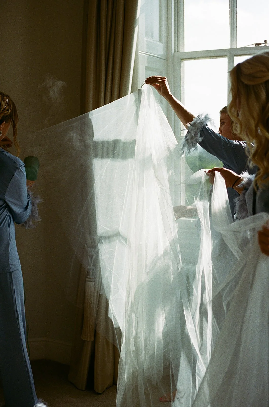 Women hanging a sheer white veil or fabric near a window with sunlight shining through.